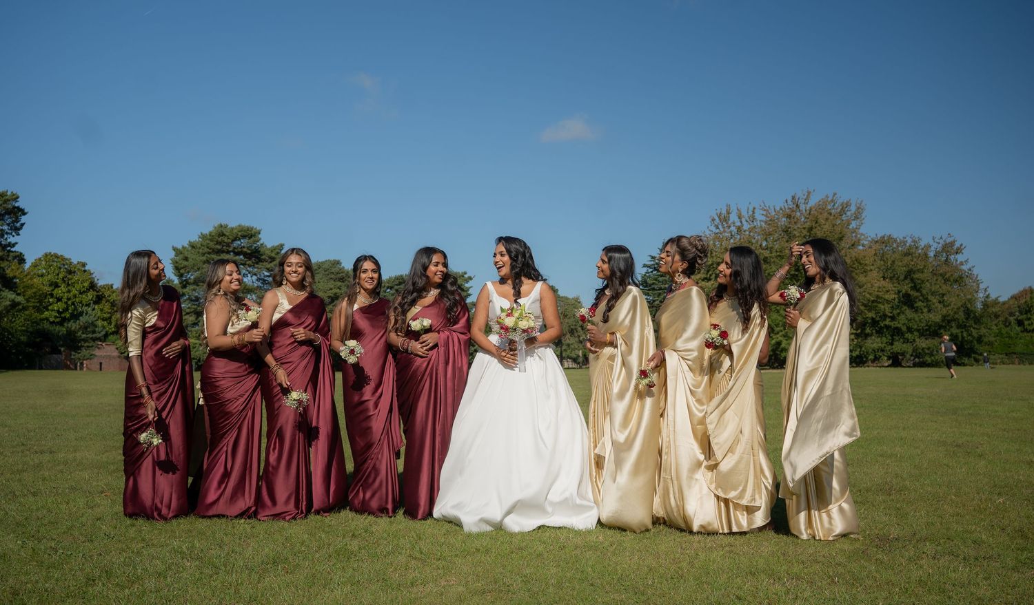 Bride in white gown stands with bridesmaids in burgundy dresses and gold sarees outdoors.