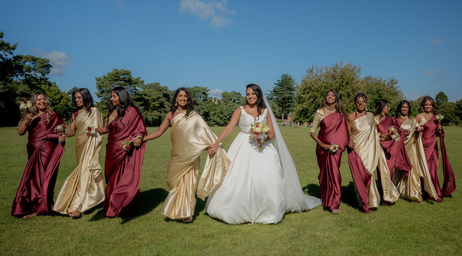 Bride holding bouquet with bridesmaids in elegant burgundy and gold dresses on lush green lawn.