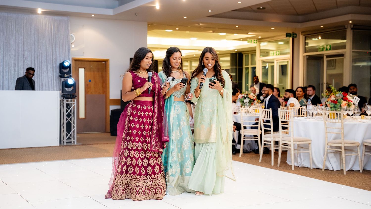 Three women in vibrant Indian lehengas stand on a dance floor holding microphones at a wedding reception.