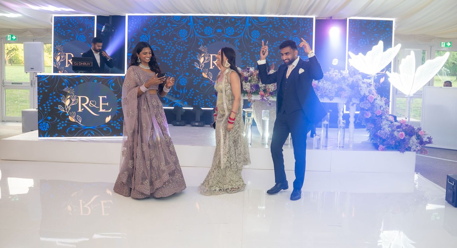 Elegant South Asian wedding reception with couple and guest celebrating on white dance floor with floral backdrop.