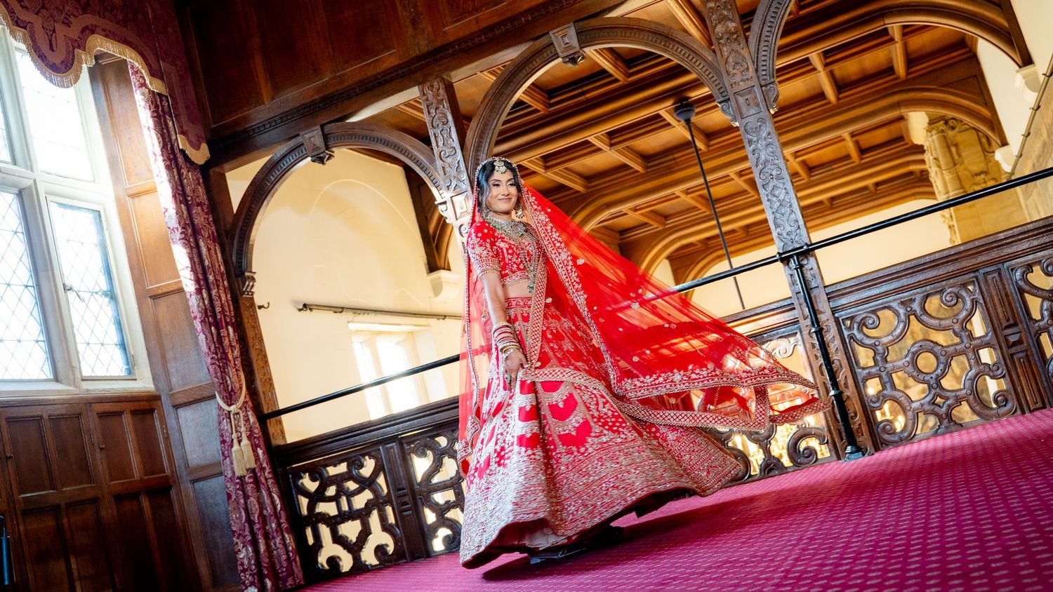 Indian bride in stunning red bridal lehenga twirling on ornate wooden staircase in elegant venue.
