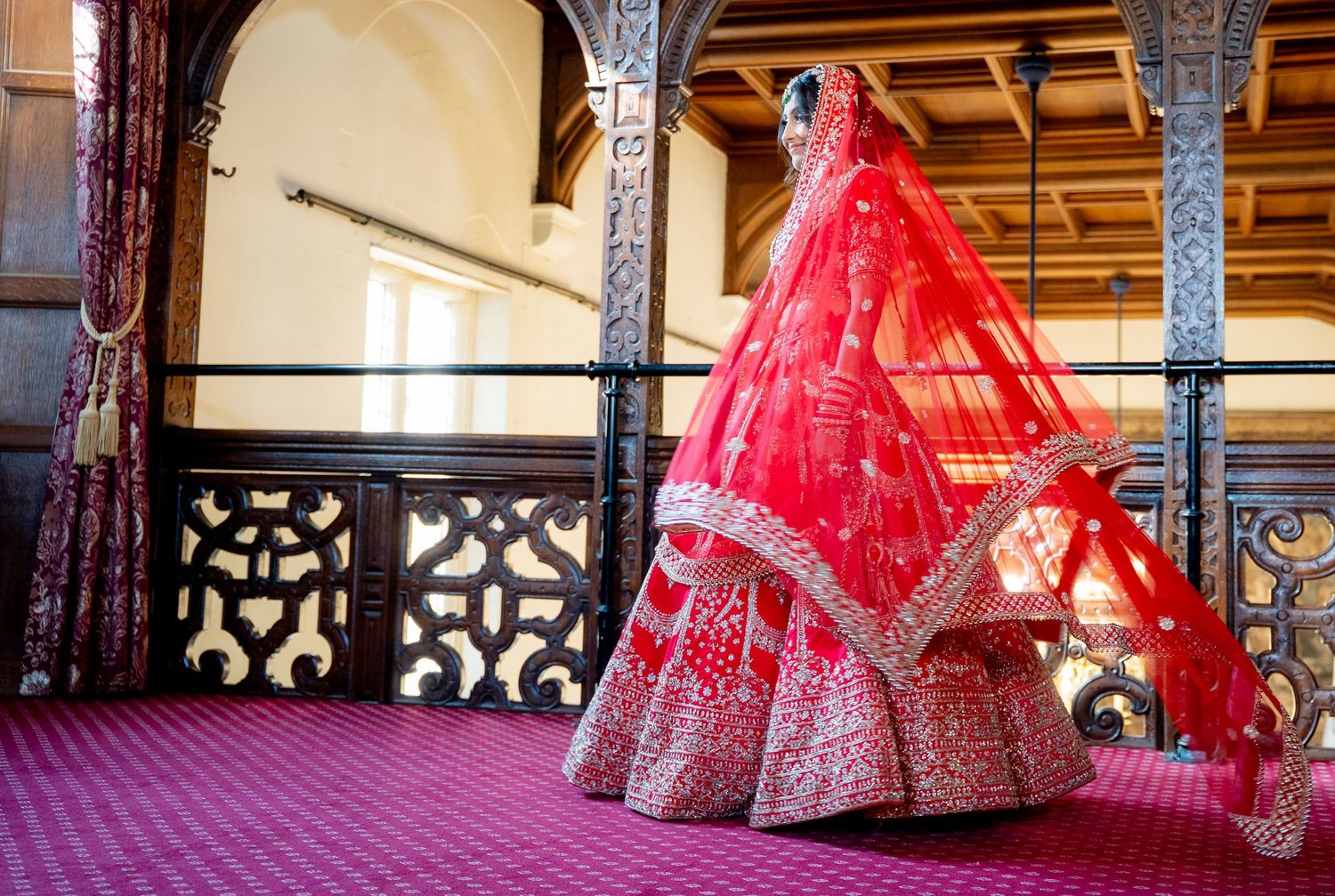 Indian bride in vibrant red lehenga with dupatta flowing on decorative balcony interior.