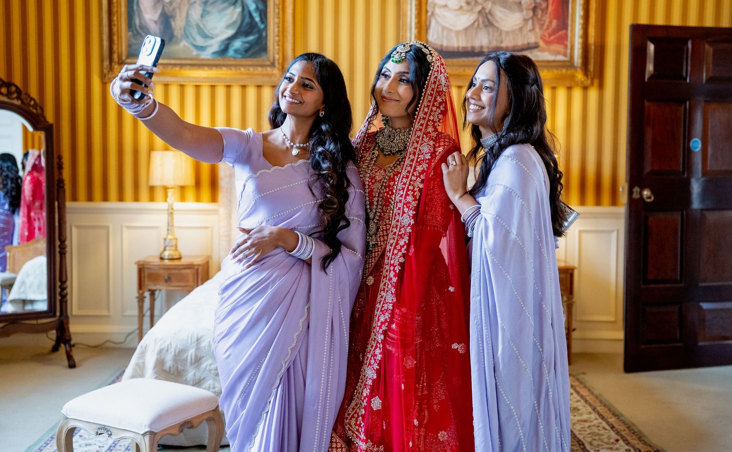 Indian bride in red bridal lehenga taking selfie with two bridesmaids in lavender sarees.