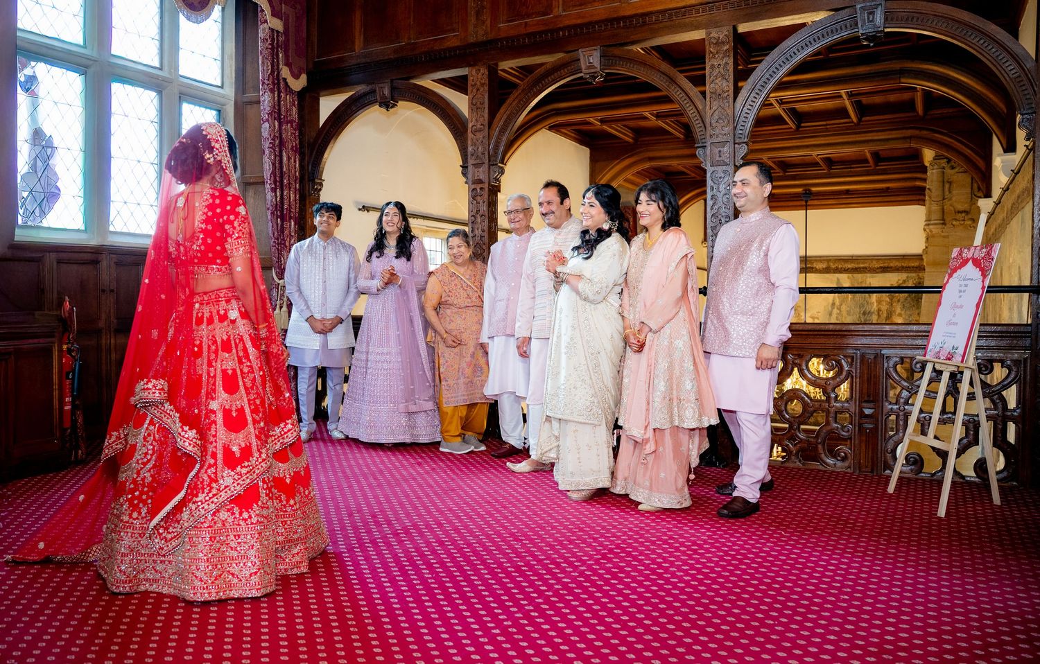 Indian bride in red lehenga greeted by family in ornate hall during wedding ceremony.