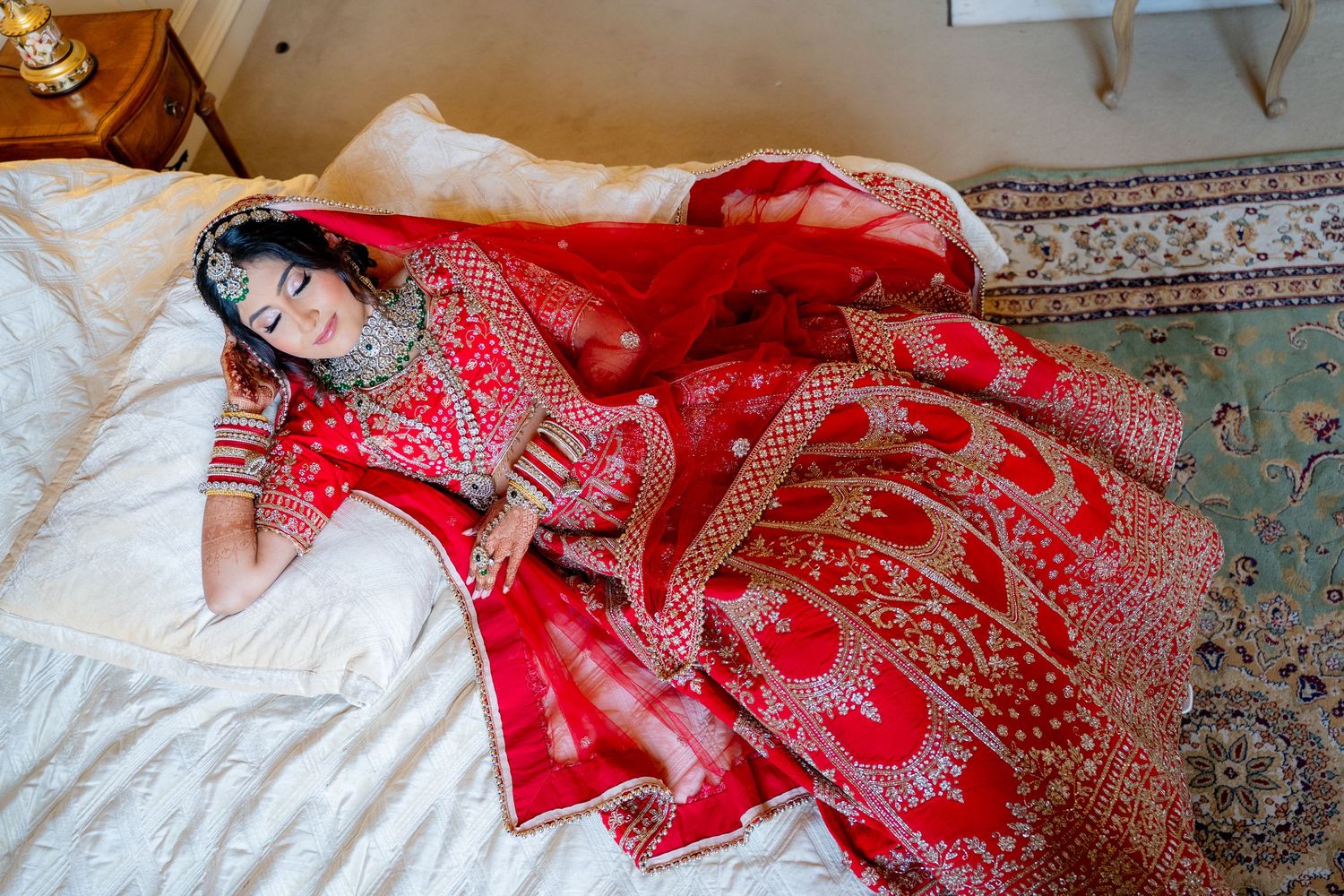 Indian bride in red embroidered lehenga lying on white bed during bridal preparation.
