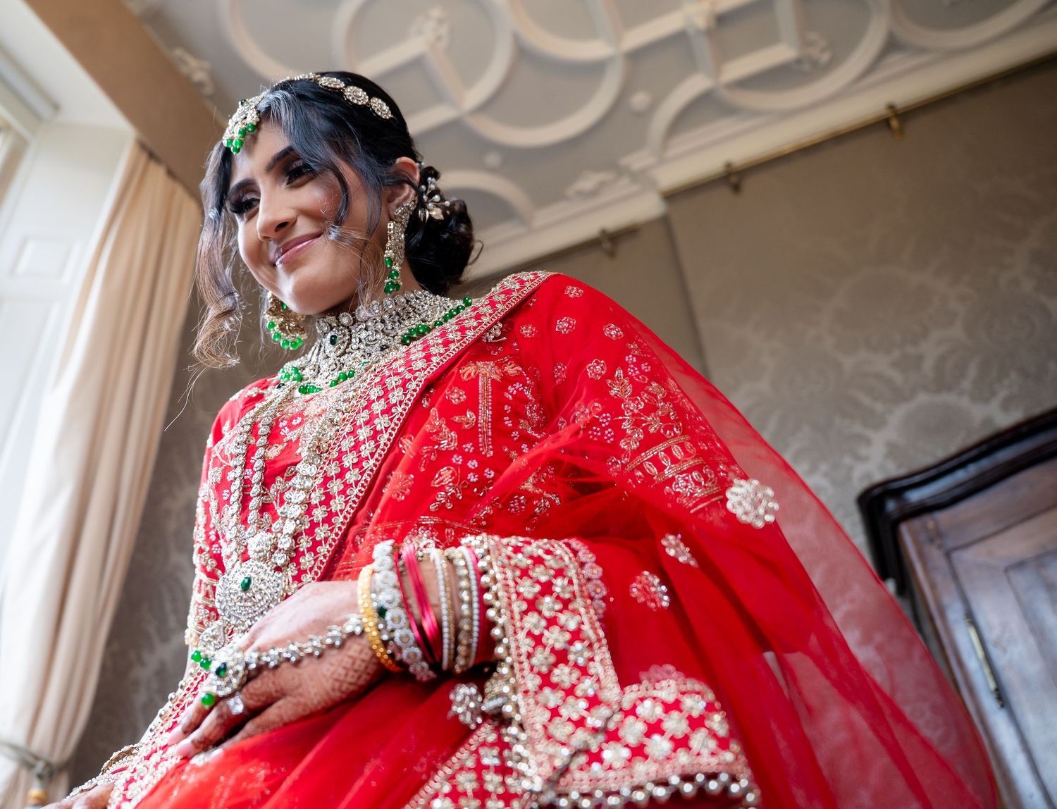 Smiling Indian bride in red lehenga with green and white jewelry in decorated room.