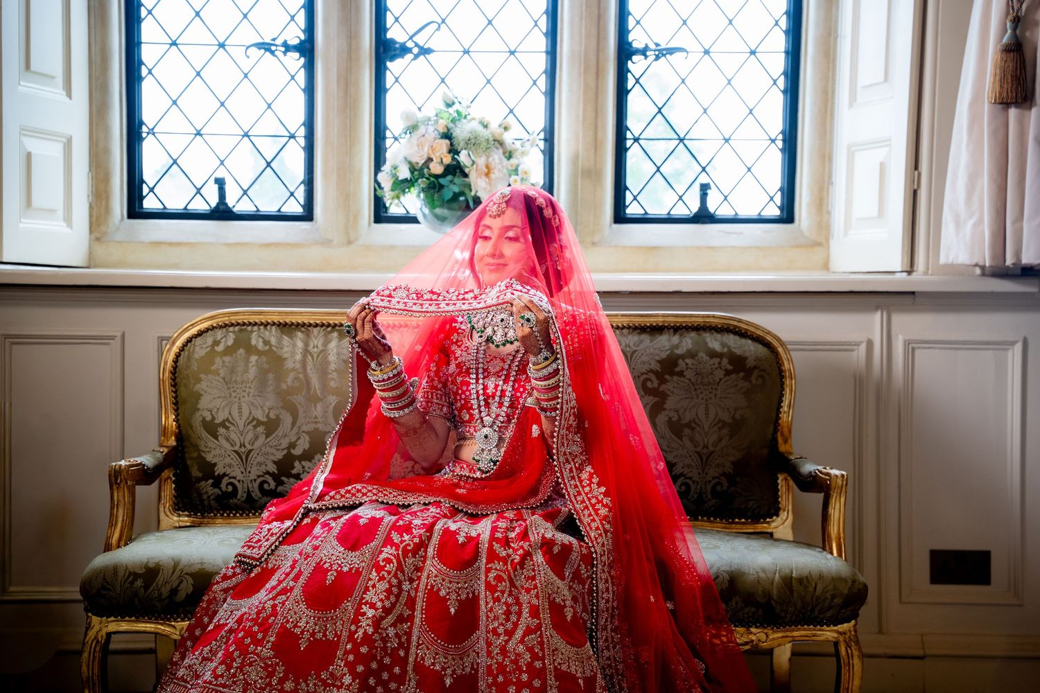 A bride in a stunning red embroidered lehenga and veil sits gracefully on a vintage sofa by a latticed window.