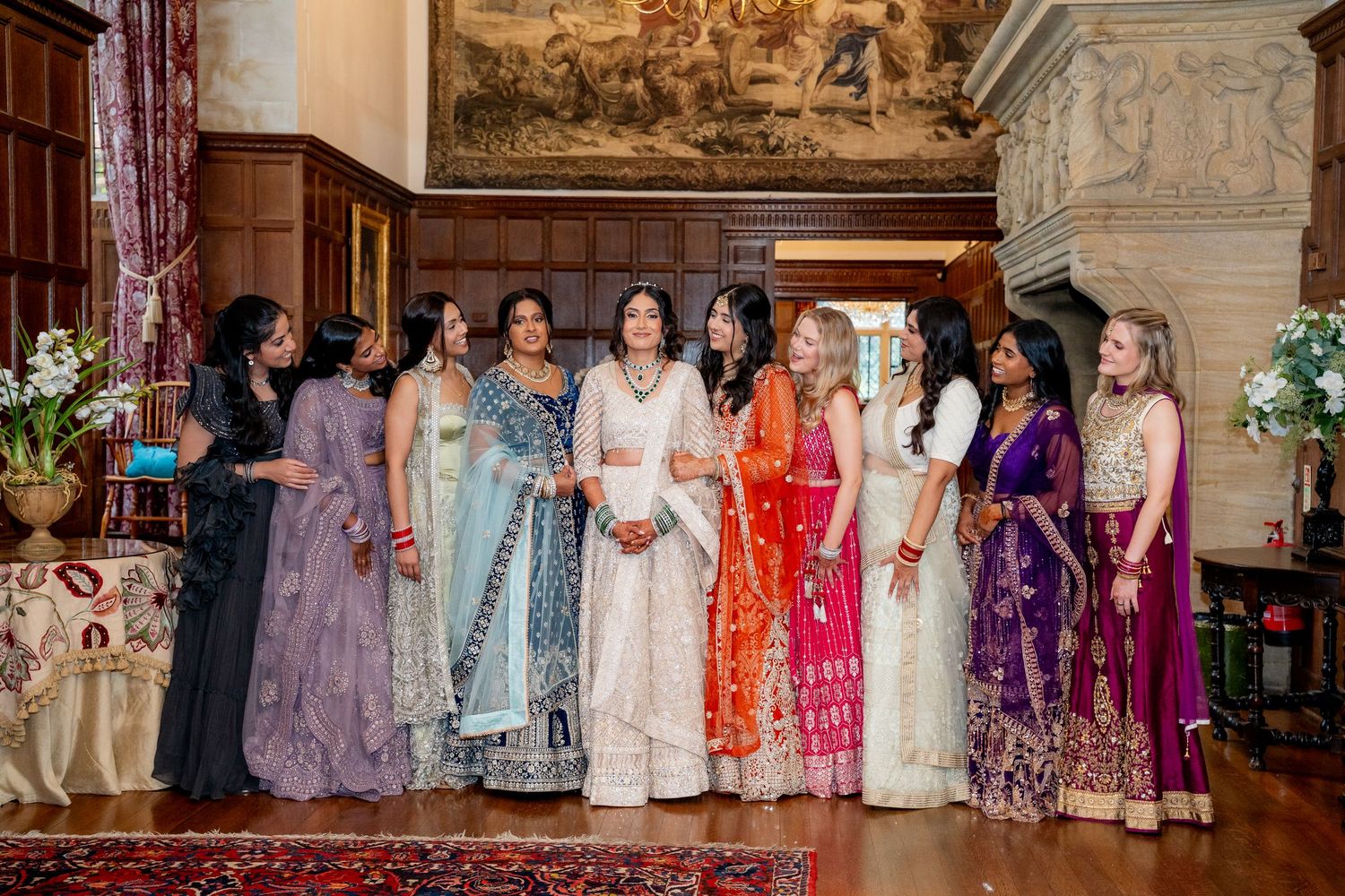 Group of women in colorful Indian wedding attire standing together in a grand manor hall.