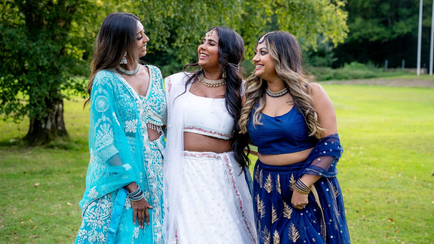 Three women in vibrant Indian attire smile together outdoors, wearing blue, white, and navy lehengas in a lush green setting.