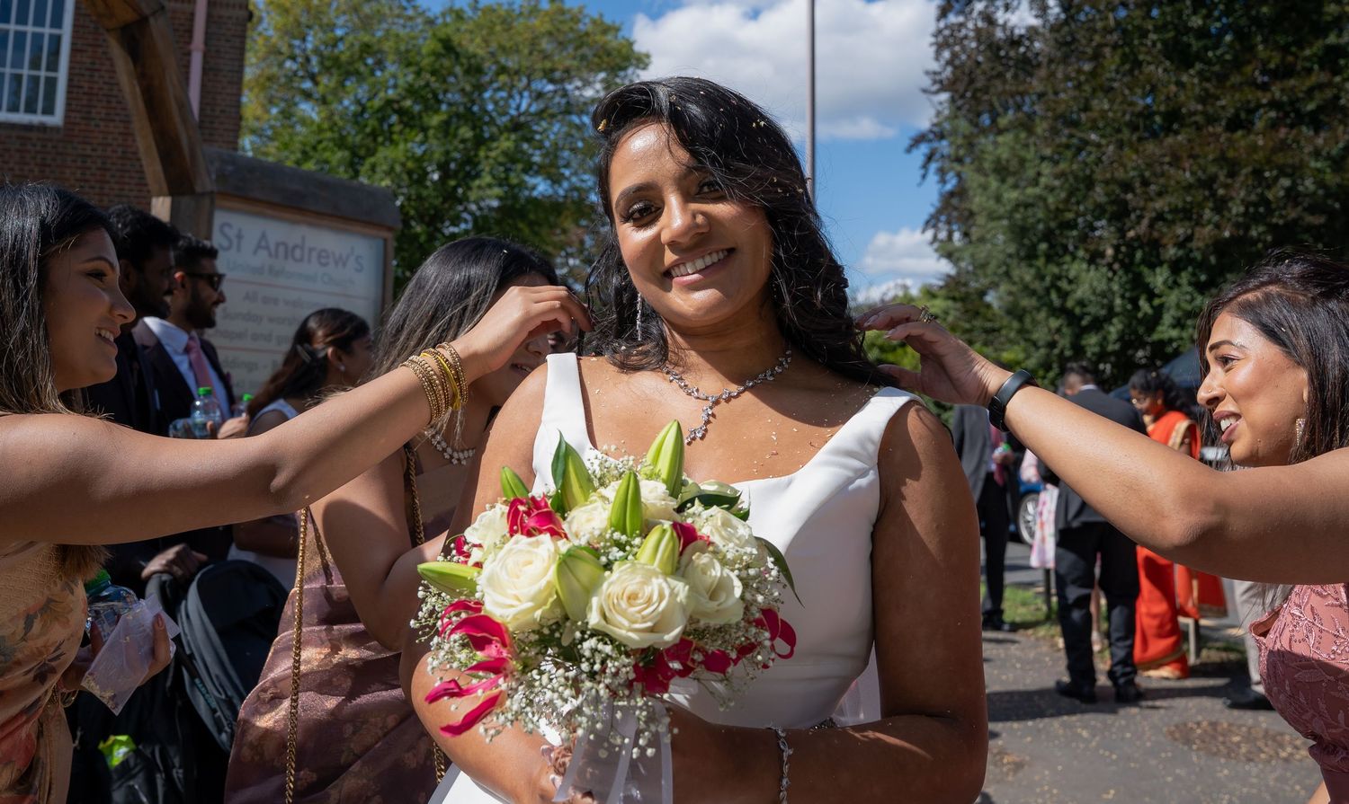 Smiling bride in white dress holding bouquet of white roses surrounded by guests outdoors on sunny day.