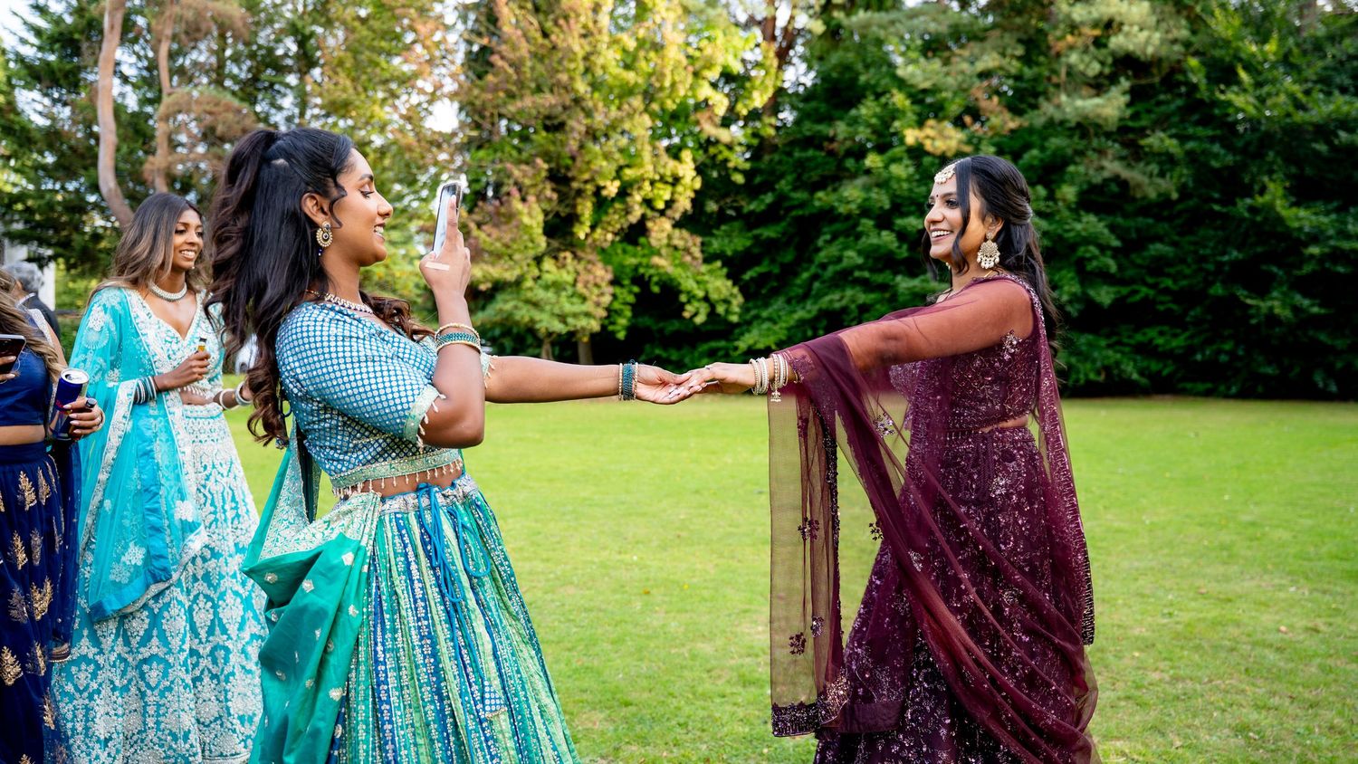 Two women in vibrant Indian lehengas dancing together joyfully on a green lawn at an outdoor wedding.
