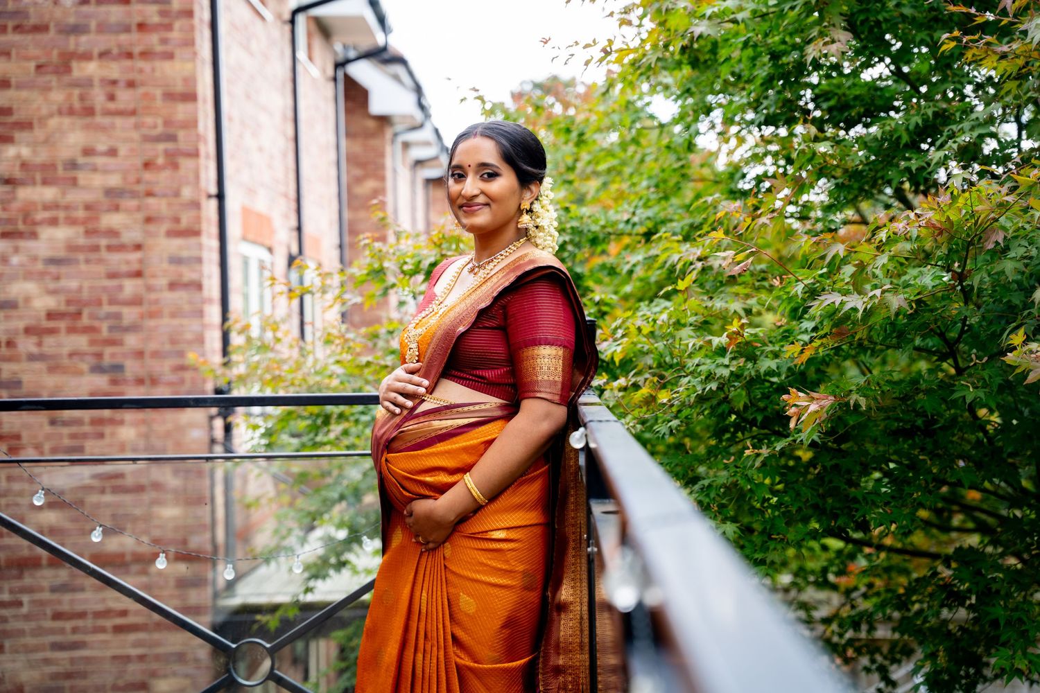 Pregnant woman in orange and red saree standing on balcony with green trees background.