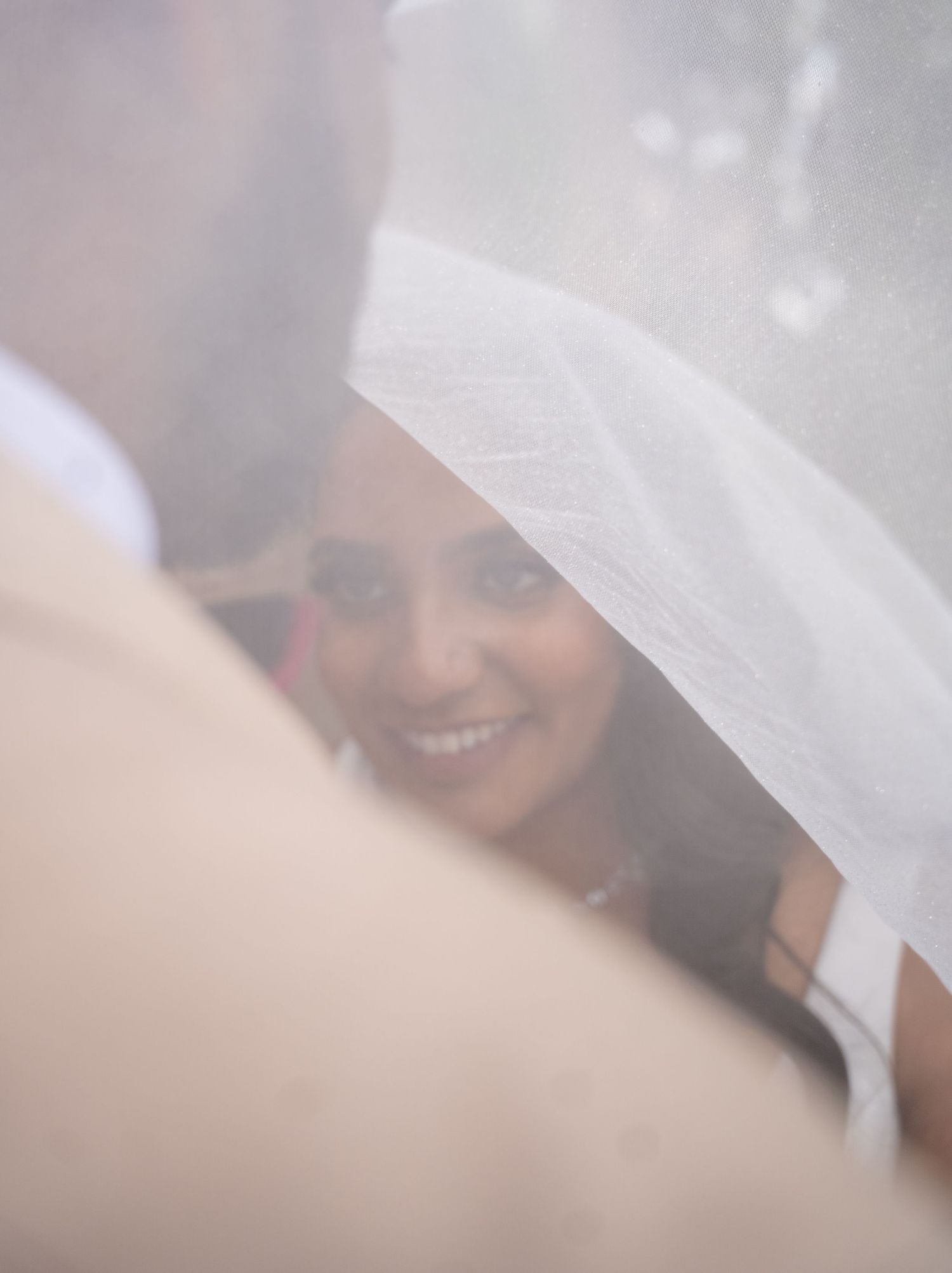 Smiling South Asian bride seen through white wedding veil looking up at groom.