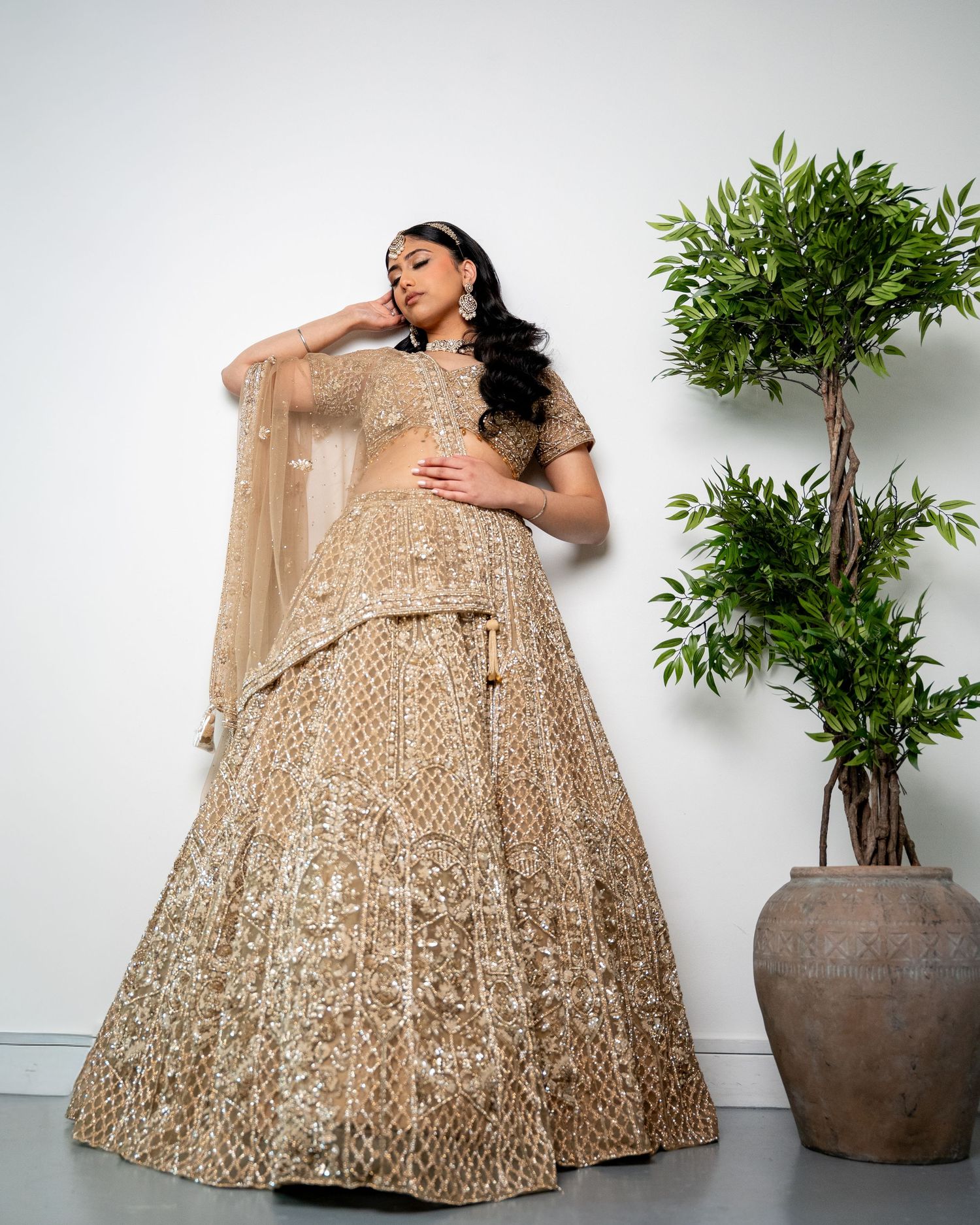 Model poses in elegant gold embroidered lehenga beside potted plant on white studio background.