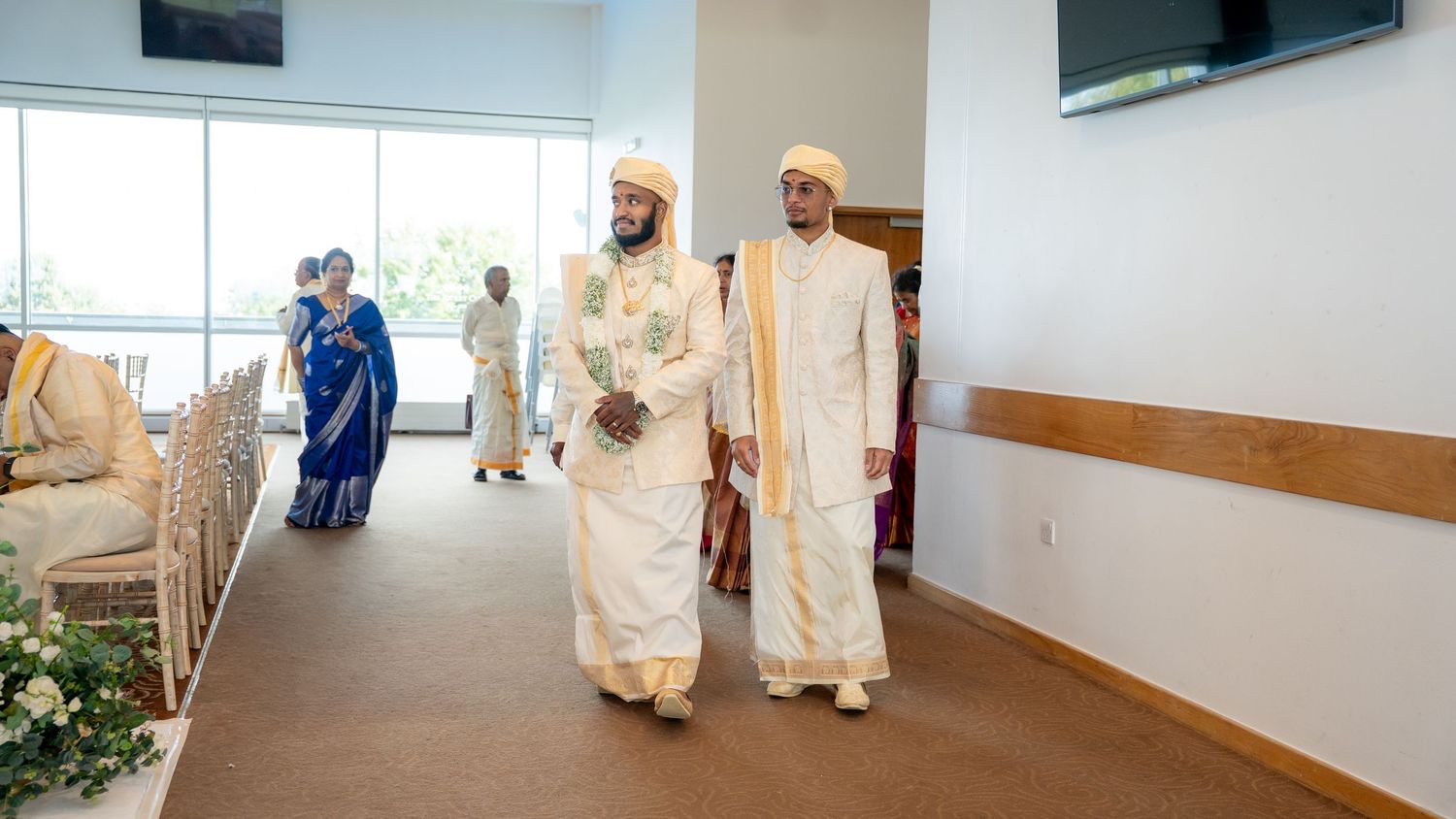 Two grooms in white traditional South Indian wedding attire walking down the aisle together.