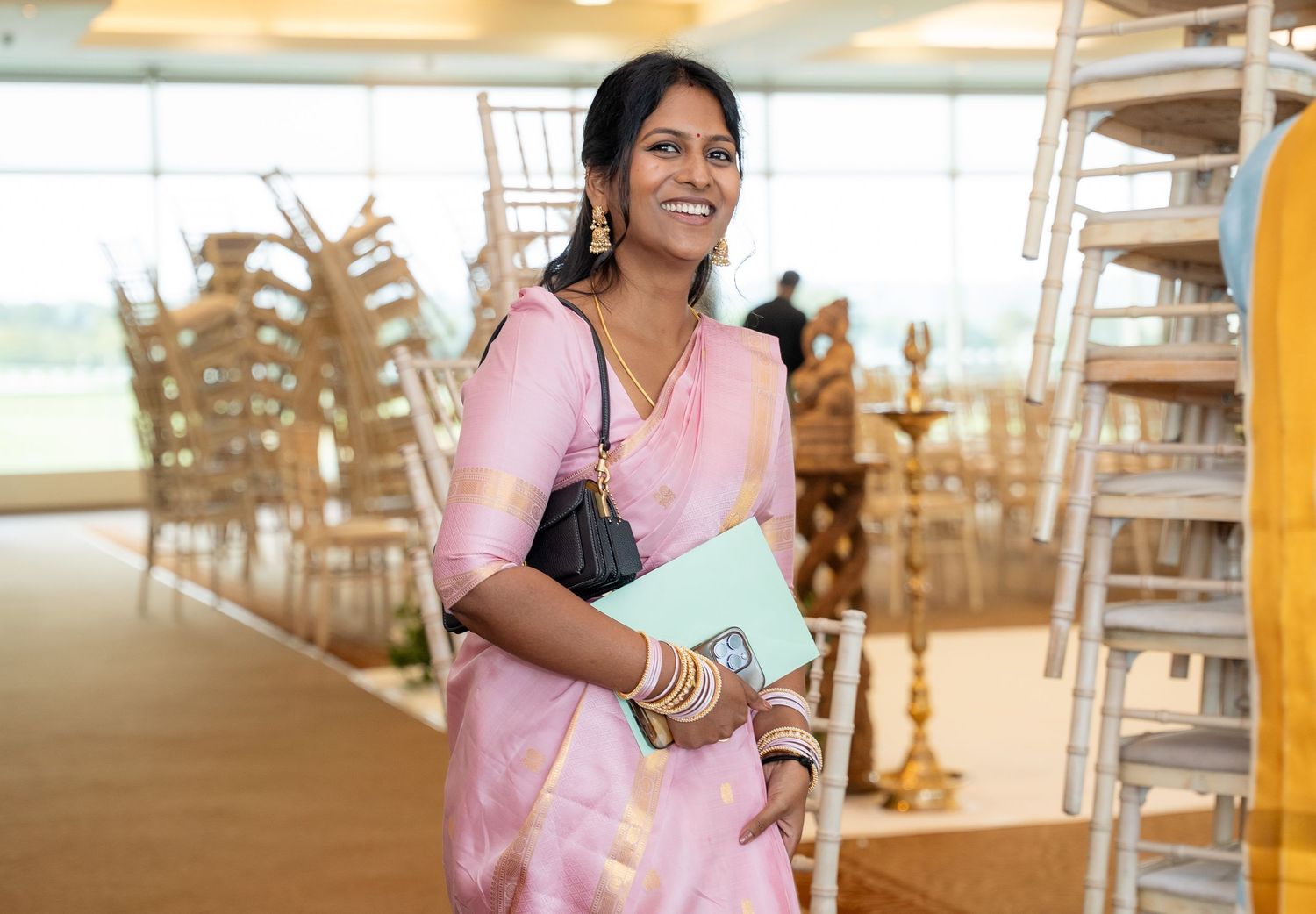 Woman in pink saree holding a black bag and blue folder smiling at an elegant wedding venue.