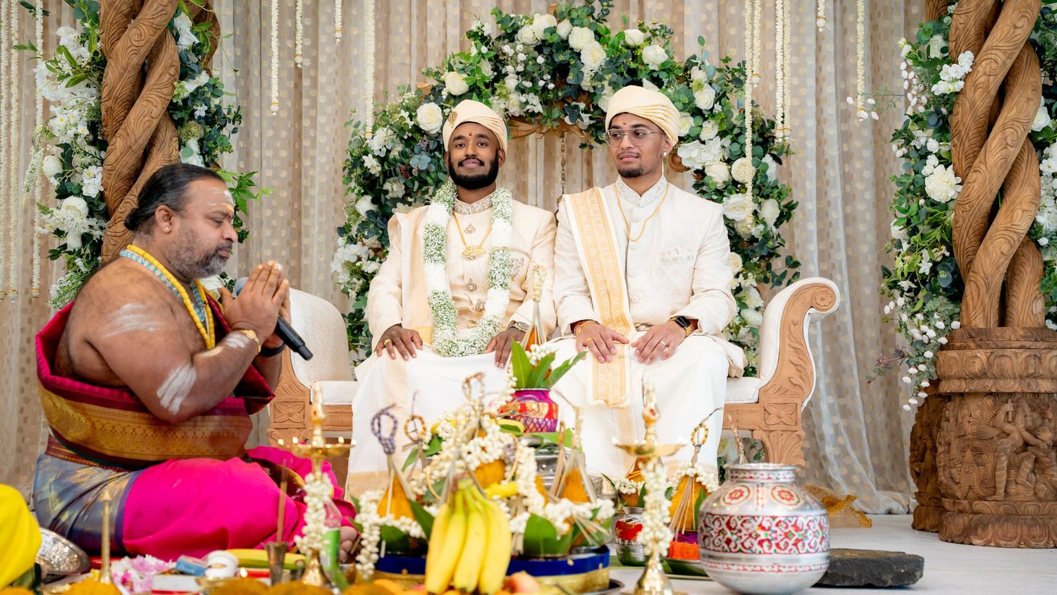 Two grooms seated at a Hindu wedding as a priest offers blessings with hands joined in prayer.