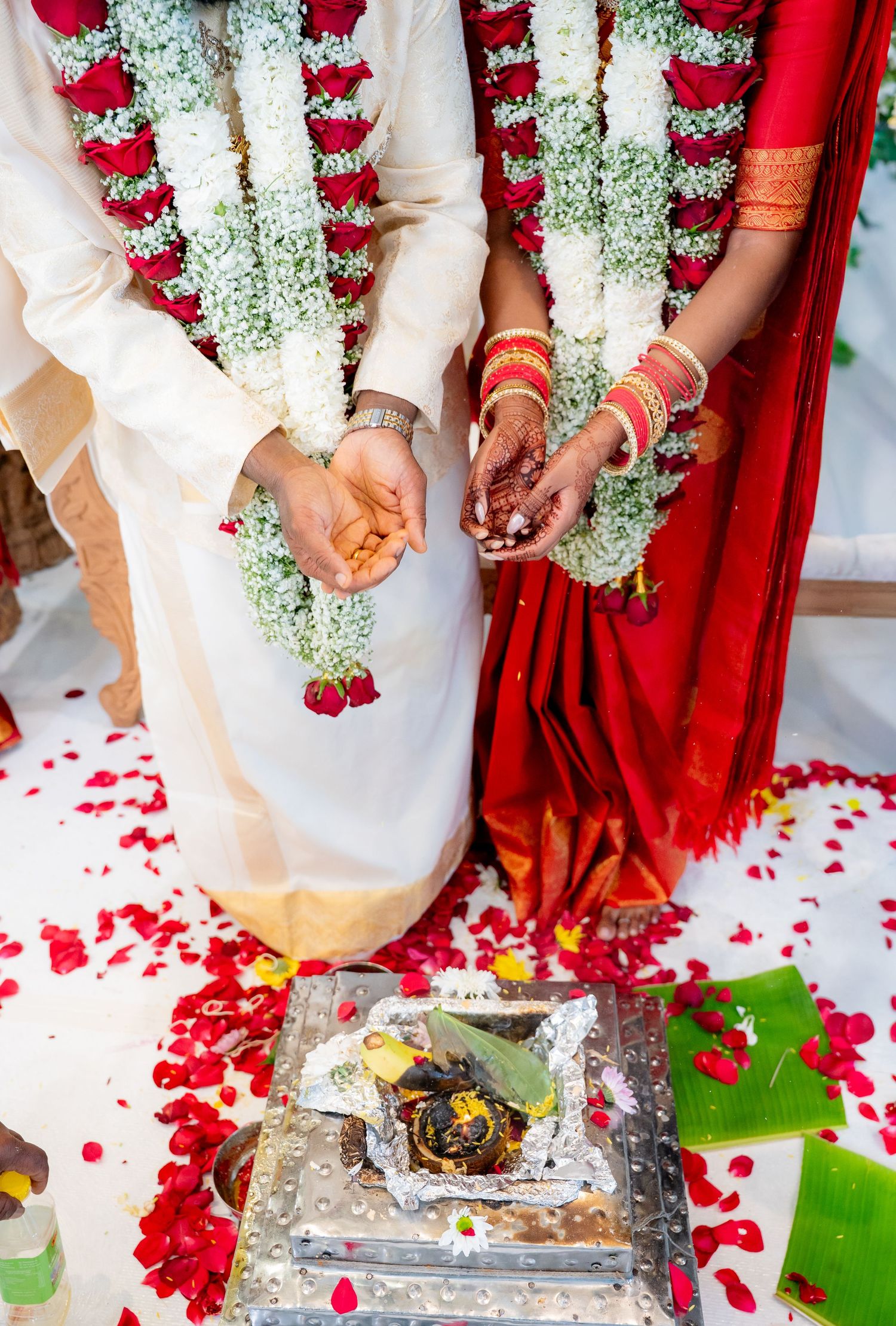 Hindu wedding couple kneeling during ritual ceremony with flower garlands and red rose petals on white floor.