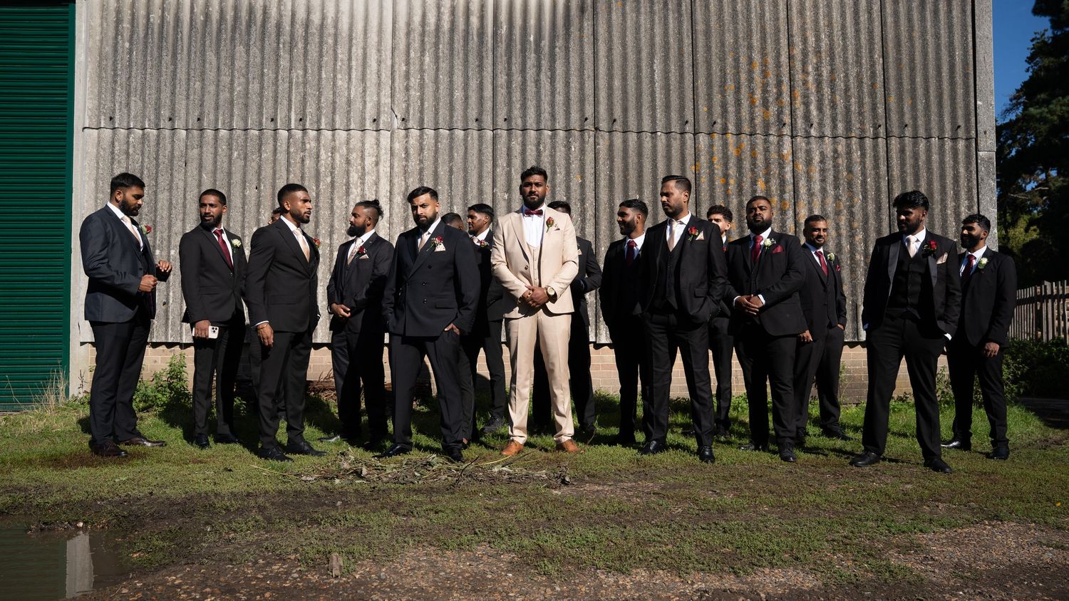 Groom in cream suit poses casually with groomsmen in dark suits outside a metal building.