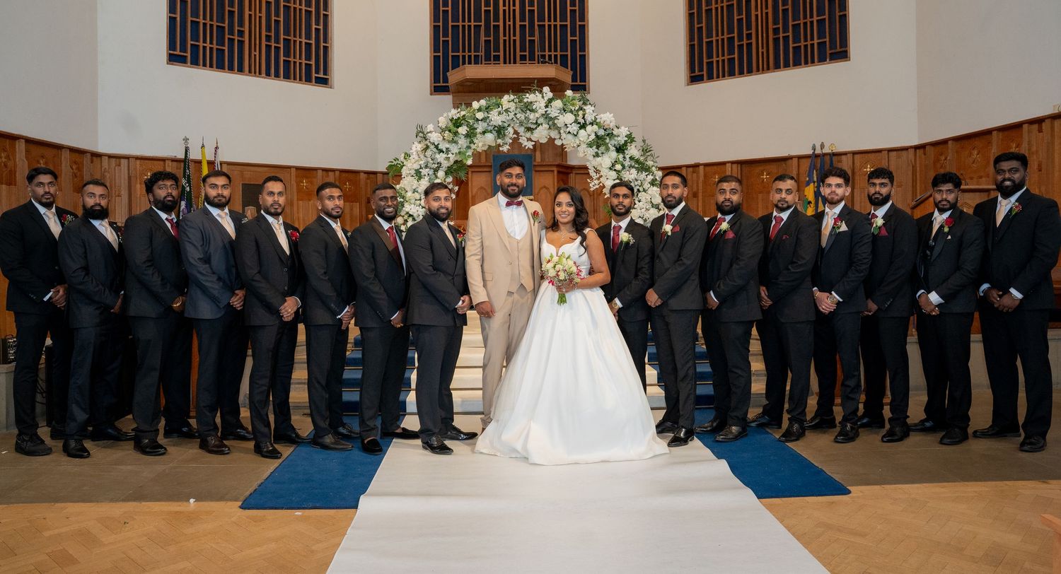 Bride and groom stand at church altar surrounded by groomsmen in dark suits at elegant wedding ceremony.