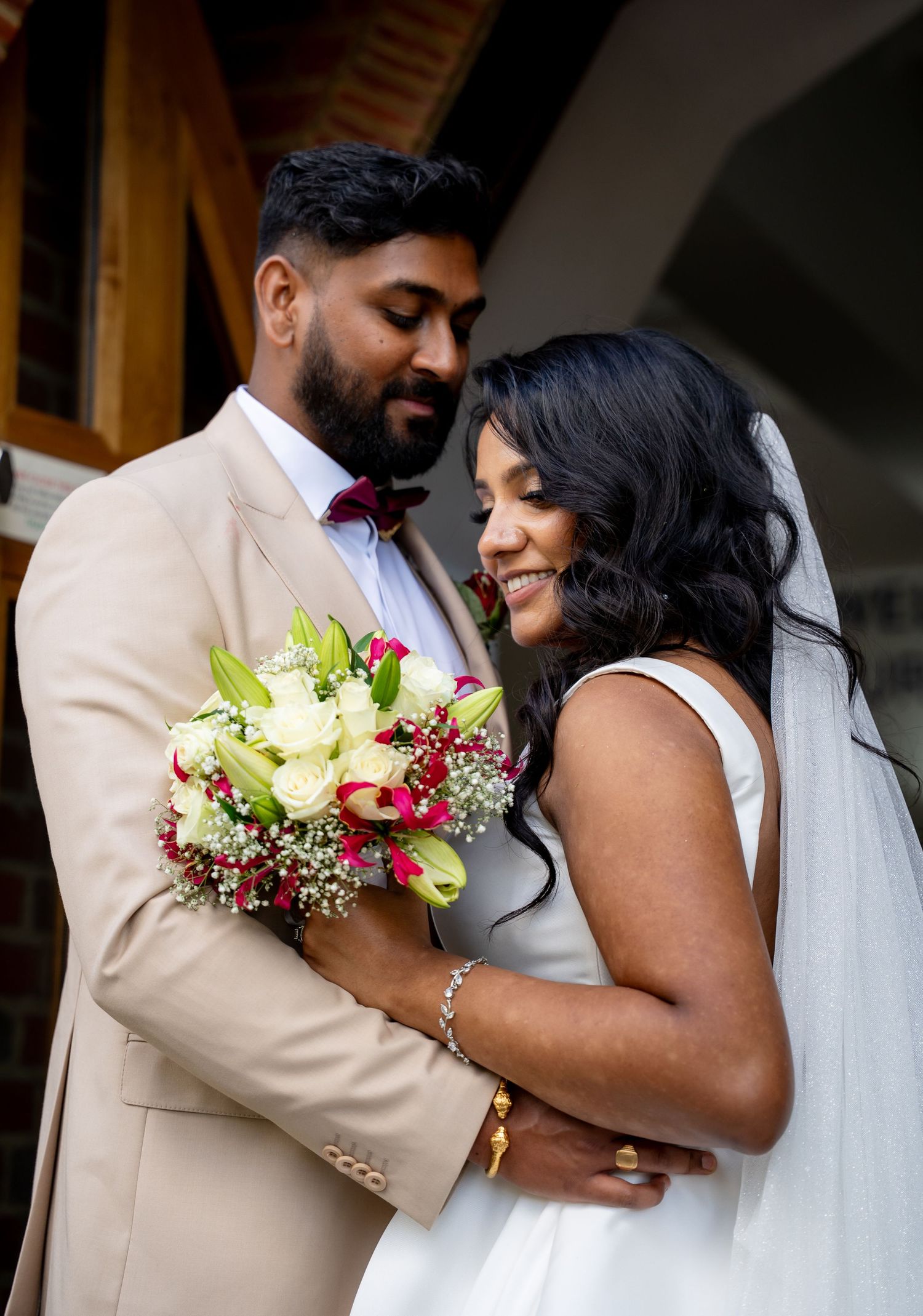 Groom in a beige suit tenderly embraces smiling bride holding a pink and white floral bouquet.
