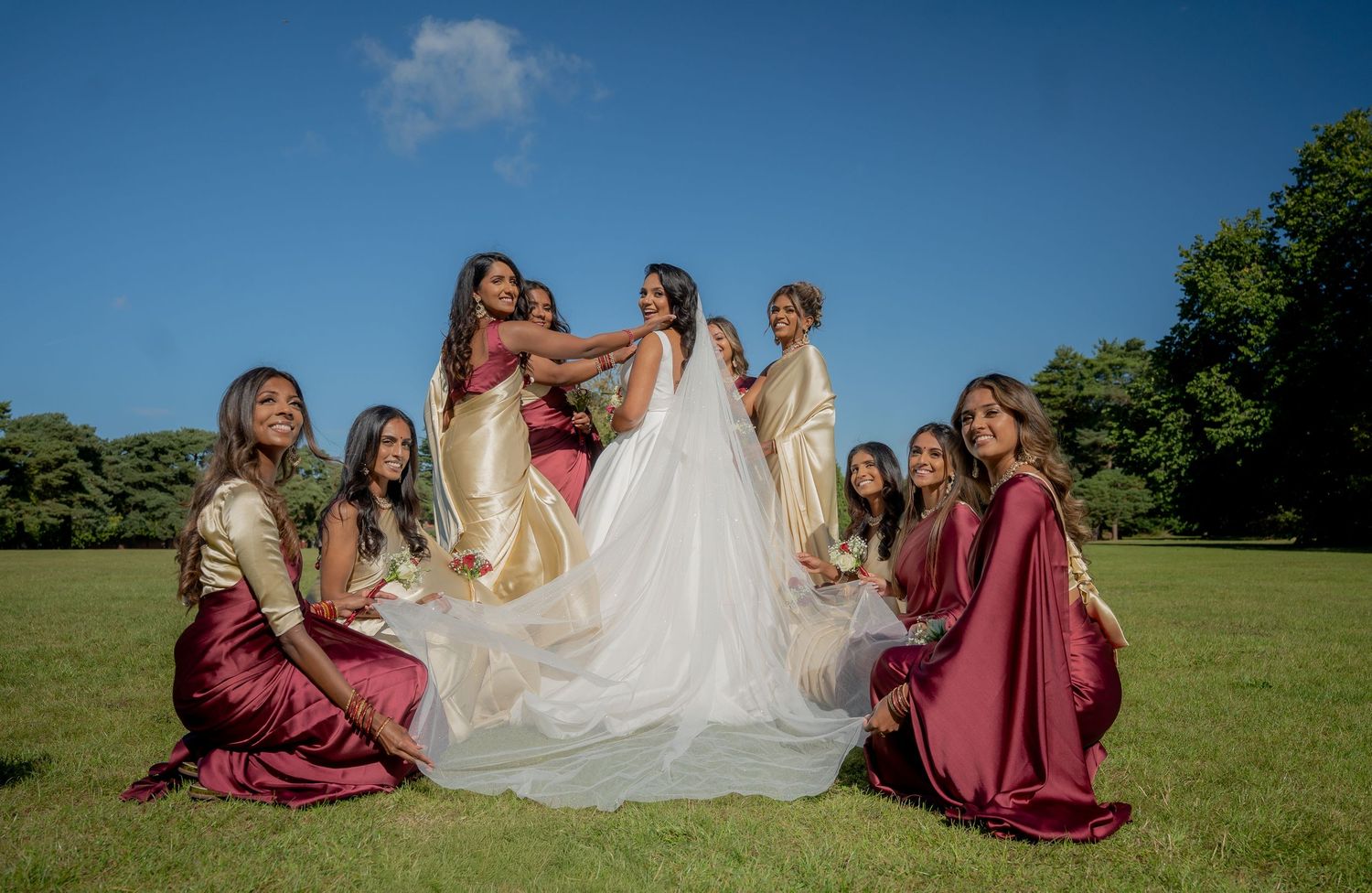 Bride in white gown surrounded by bridesmaids in gold and burgundy dresses on green lawn under blue sky.