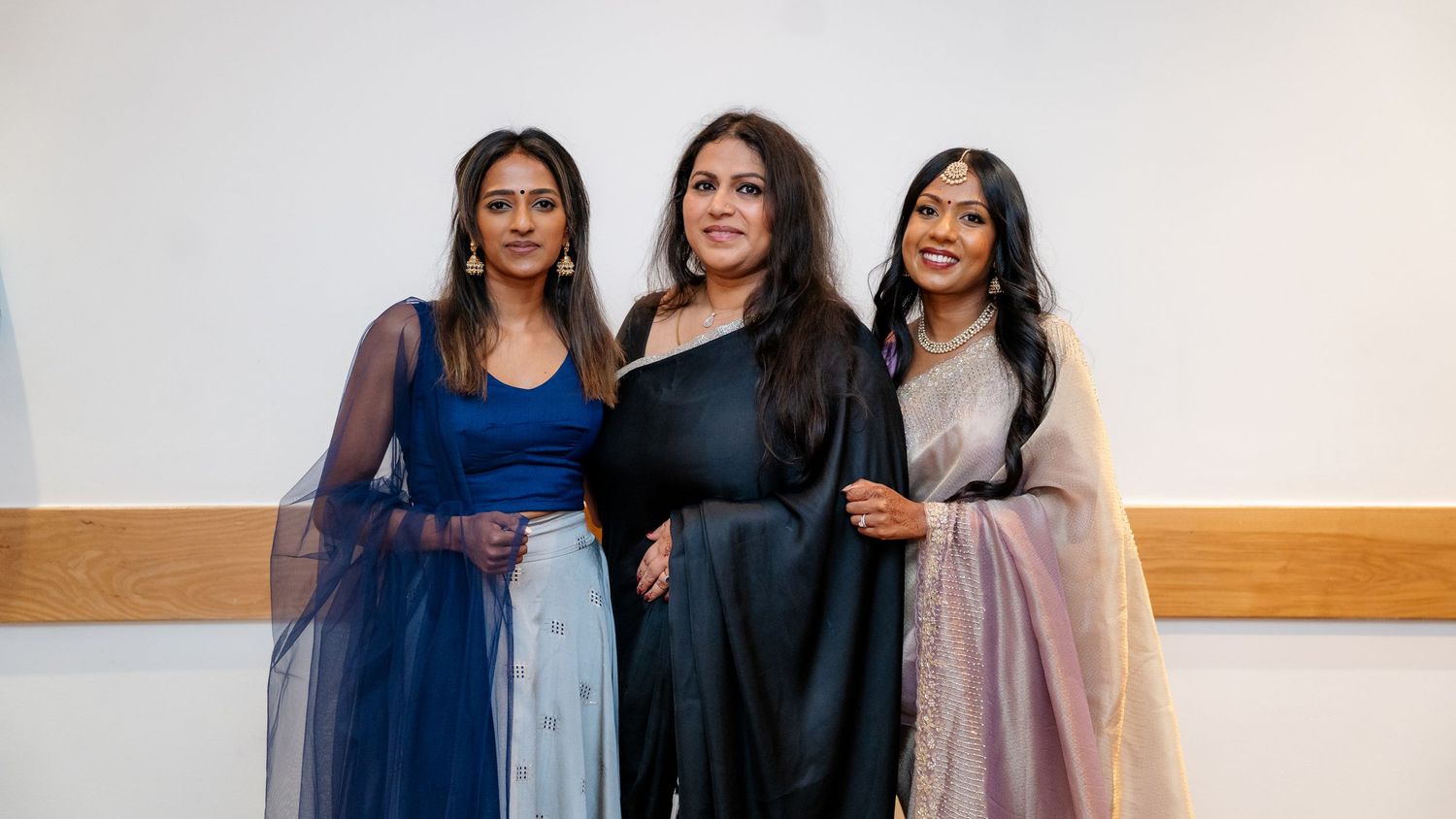 Three women dressed in traditional Indian outfits smile together at a celebration.
