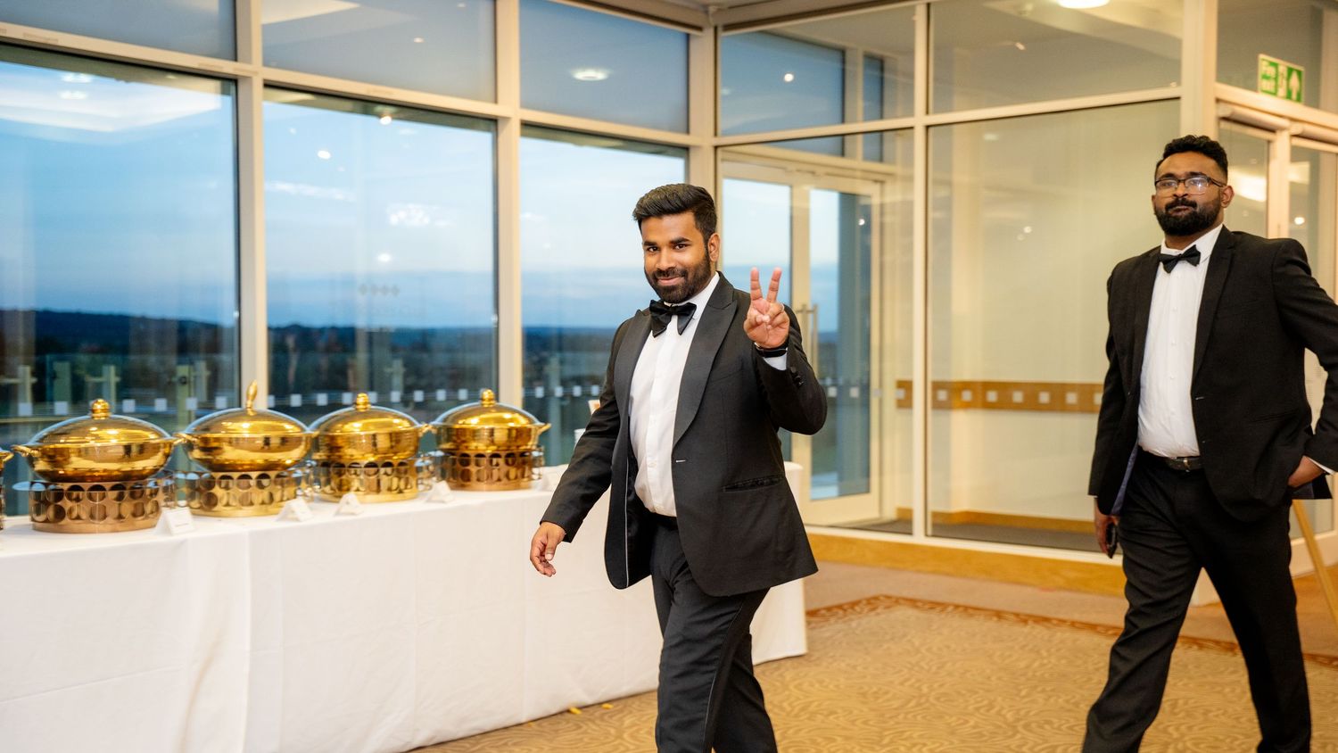 Man in tuxedo waving at a wedding reception with gold serving dishes in background.