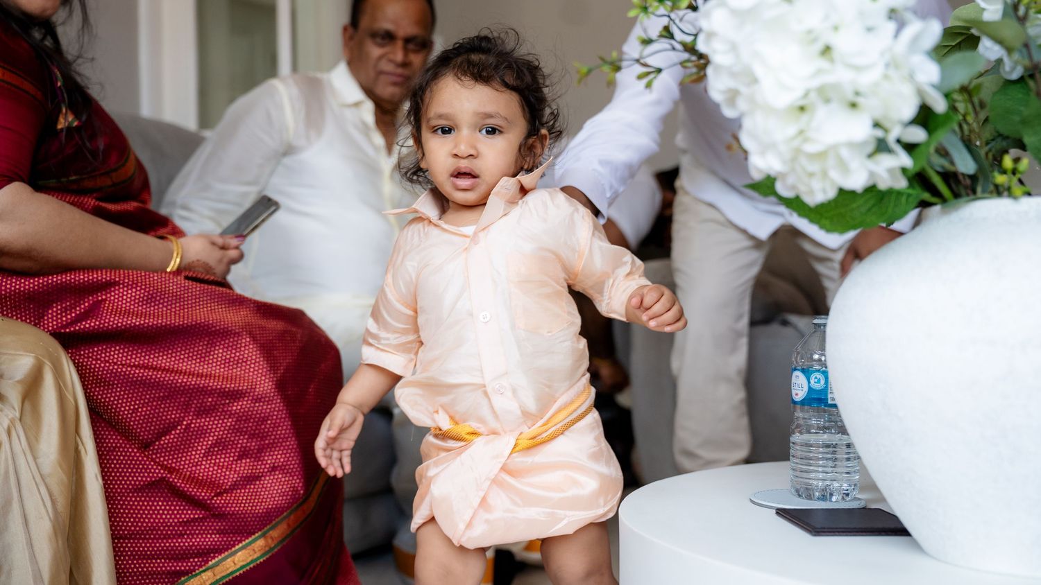 Toddler in traditional peach Indian attire walking confidently at a family wedding celebration indoors.