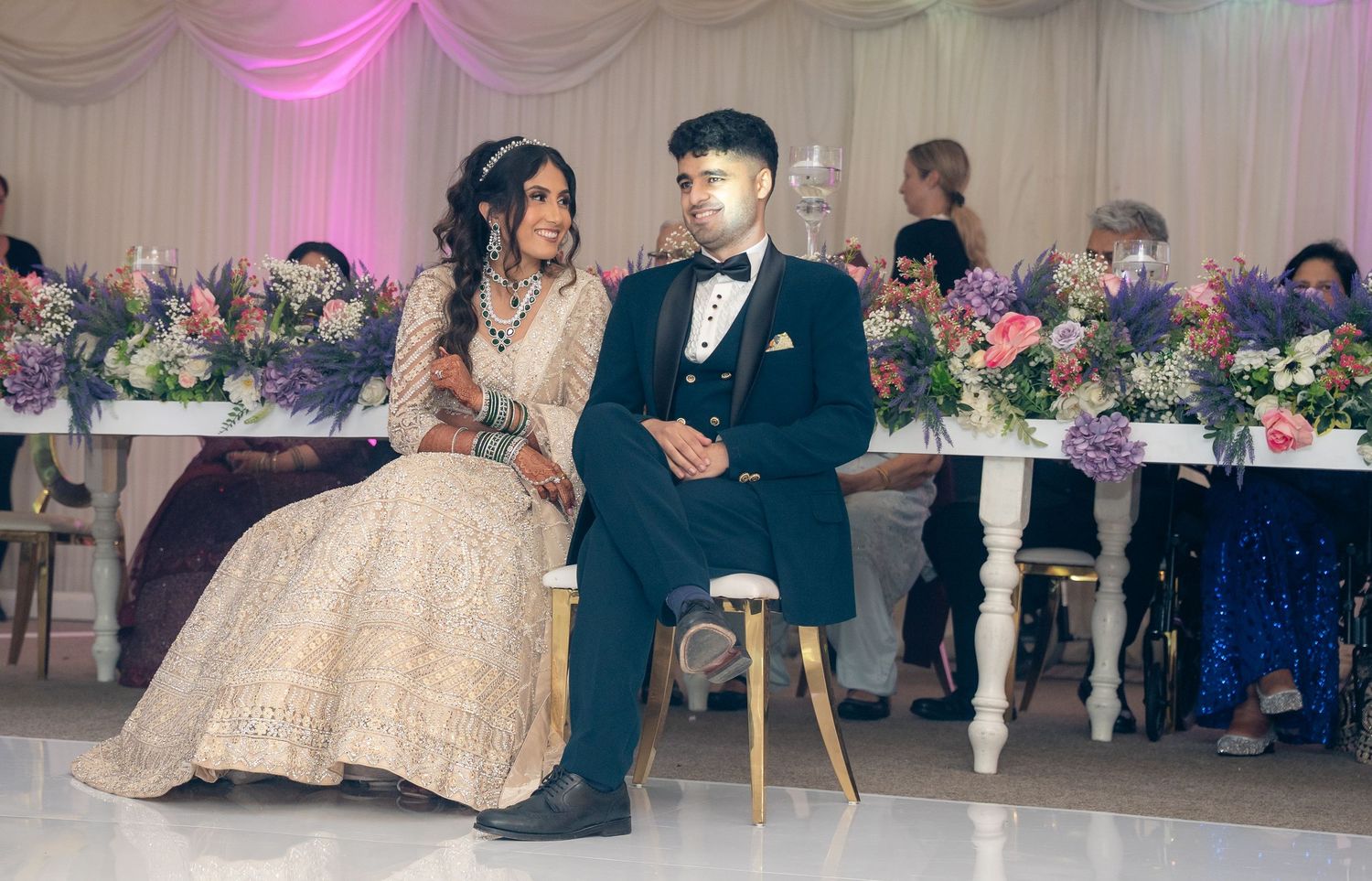 Bride and groom seated together at elegant wedding reception with floral arrangements and guests in background.