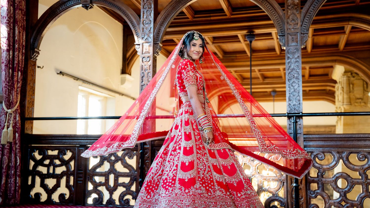 A bride in a stunning red embroidered lehenga with a flowing dupatta poses gracefully on an ornate balcony.