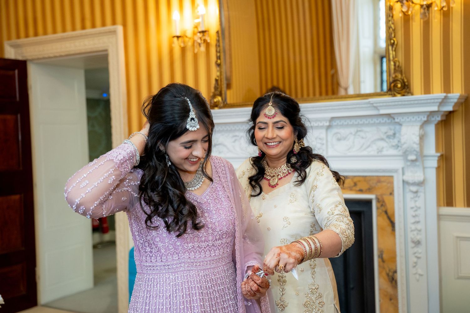 Two women in traditional Indian attire smile together near a fireplace during bridal wedding preparations.