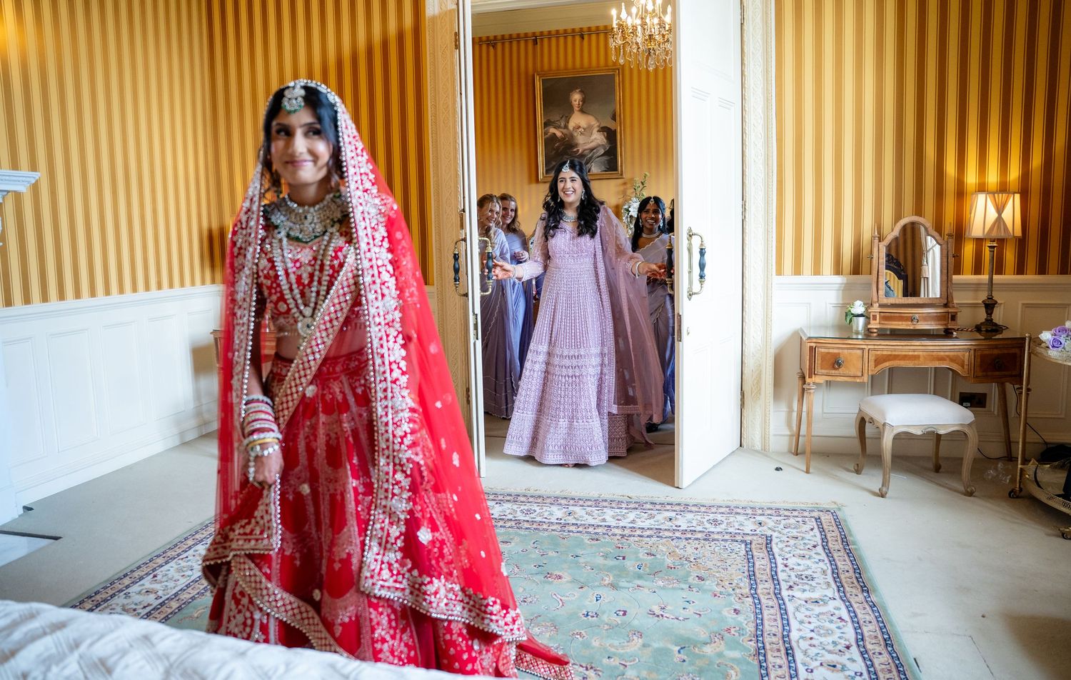 Bride in red traditional Indian bridal lehenga with gold embroidery stands in elegant room as bridesmaids look on.