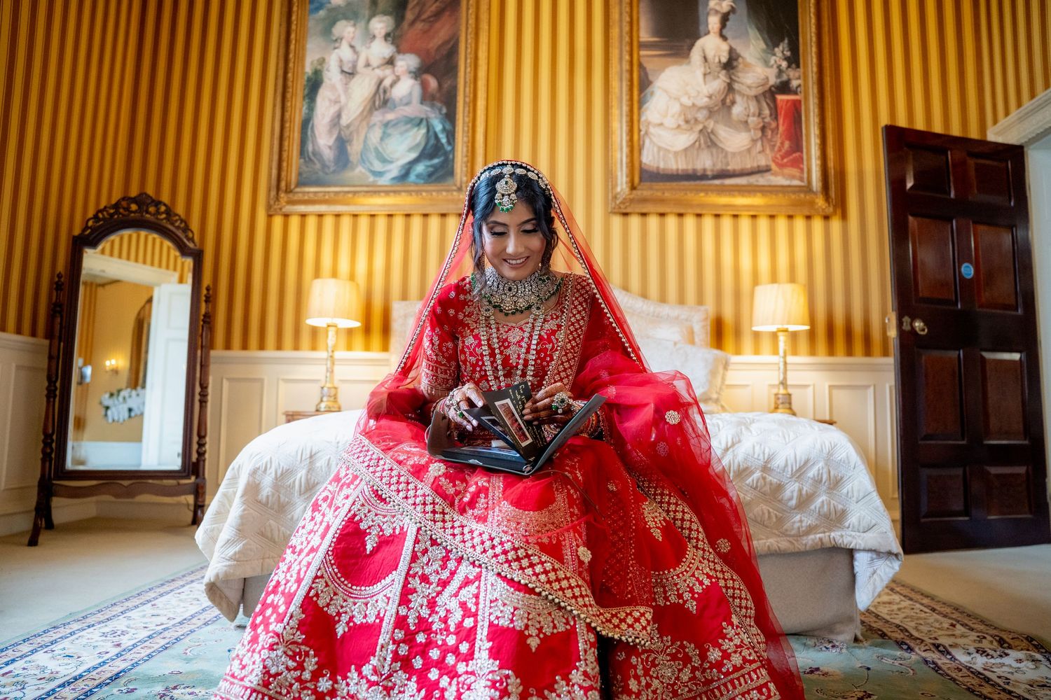Indian bride in red lehenga sitting on bed in elegant yellow room.