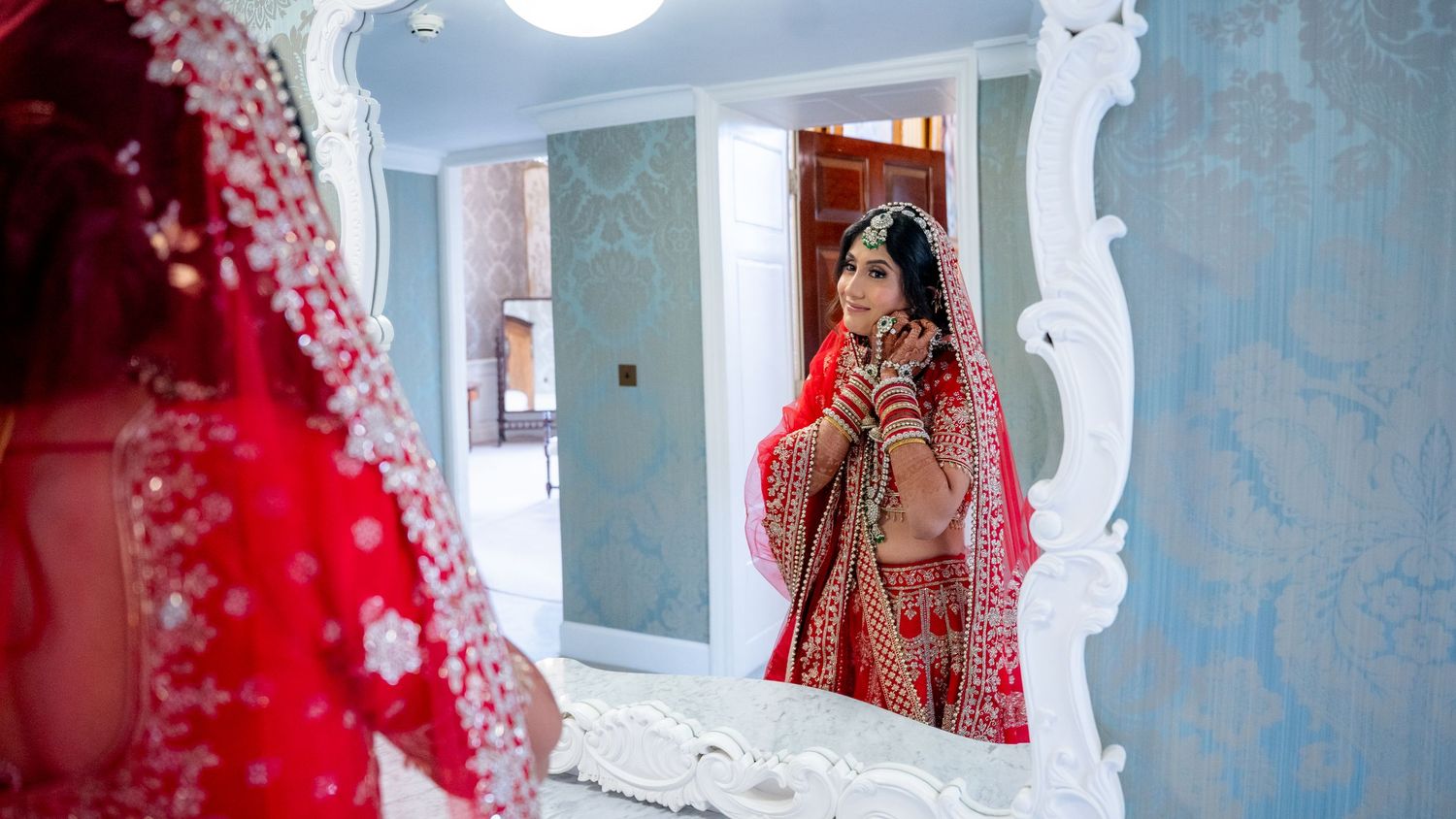 Indian bride in red lehenga adjusting jewelry while viewing reflection in decorative white mirror.