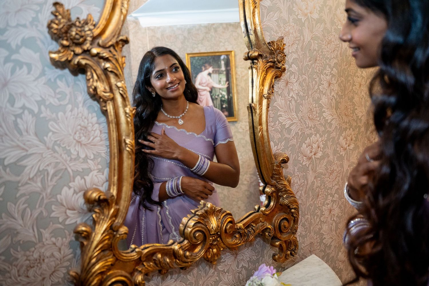 Woman in lavender saree admiring herself in ornate gold-framed mirror with floral wallpaper.