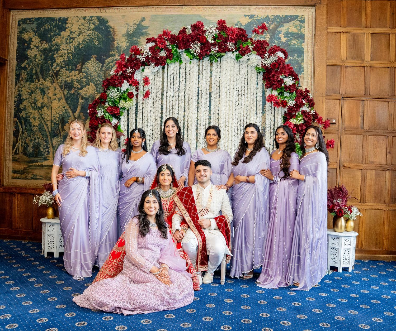 Indian bride and groom with bridesmaids in lavender sarees under floral arch.