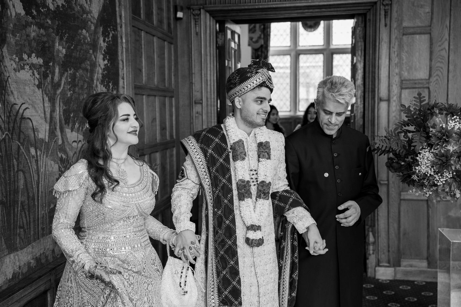 Black and white photo of a couple in traditional South Asian wedding attire walking with a guest in an elegant venue.