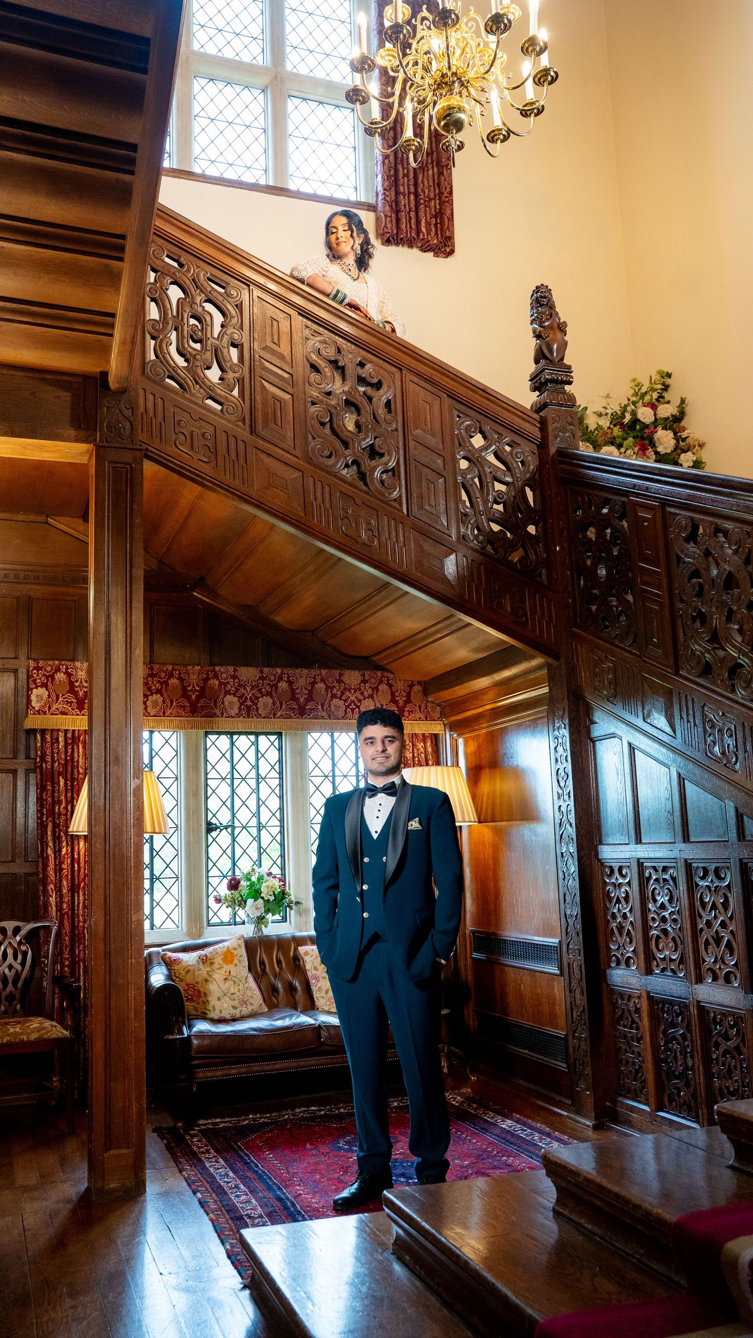 Groom in dark tuxedo stands in ornate wooden manor hall while bride looks down from carved balcony above.