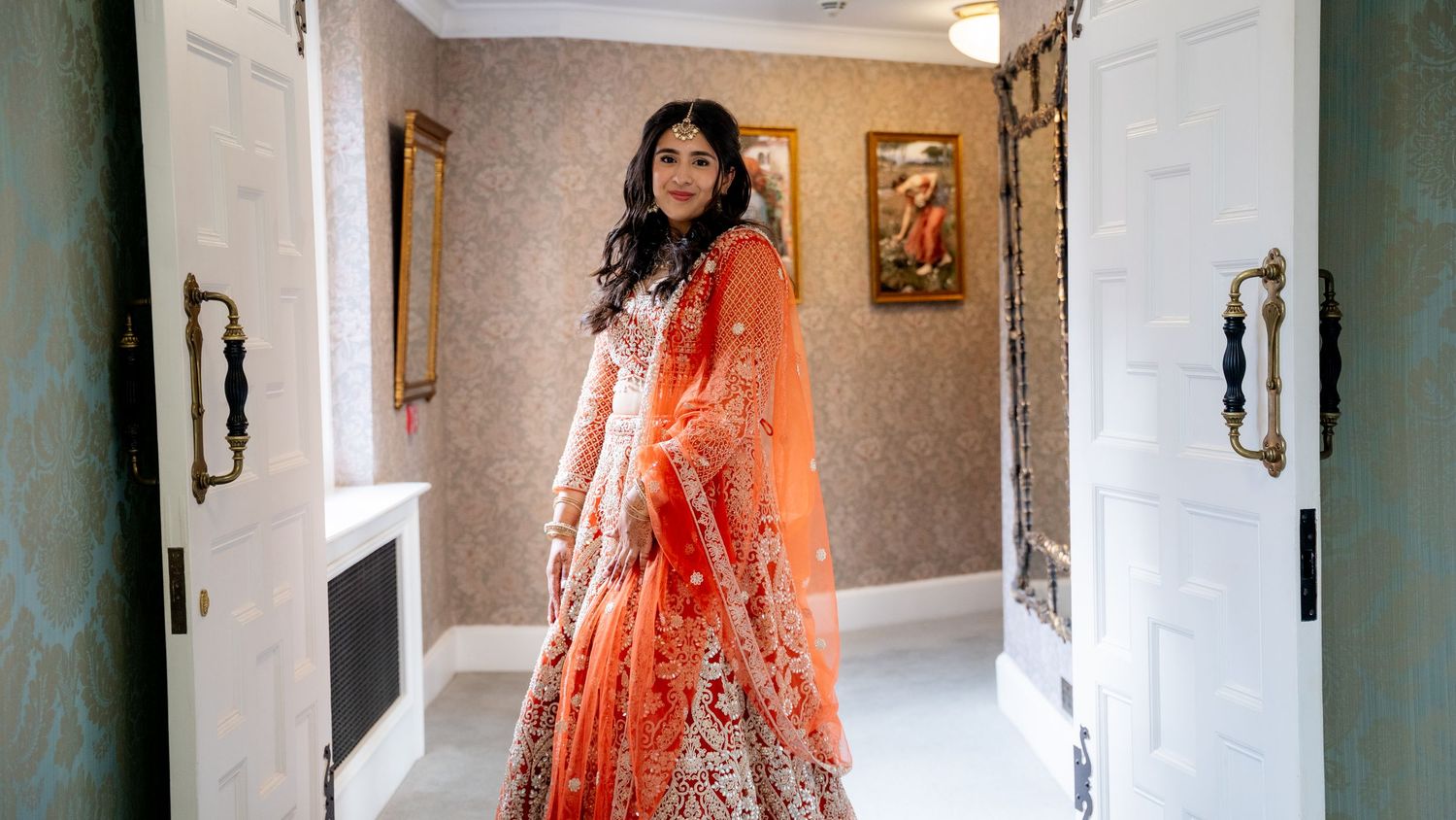 Indian bride in orange lehenga standing elegantly in a doorway of a decorated room.
