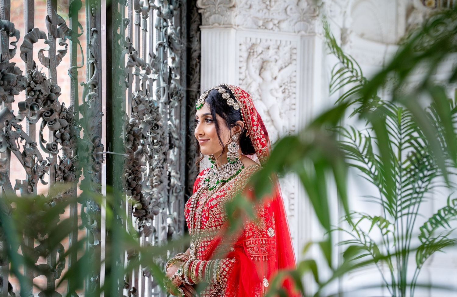 Indian bride in red lehenga with gold jewelry poses gracefully by ornate iron gates at a grand venue.
