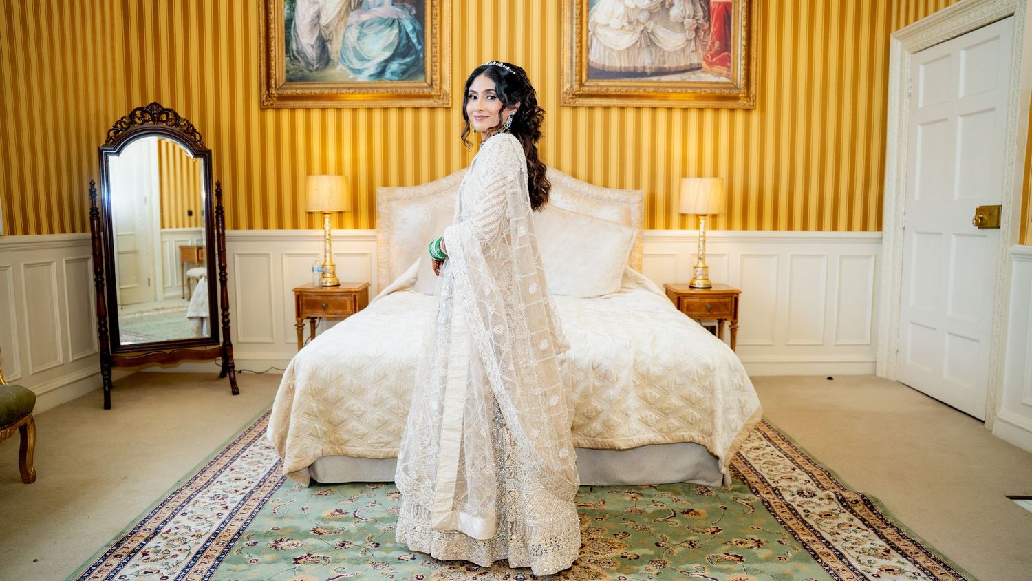 Bride in white embroidered anarkali stands elegantly in a luxurious yellow-walled hotel bedroom.