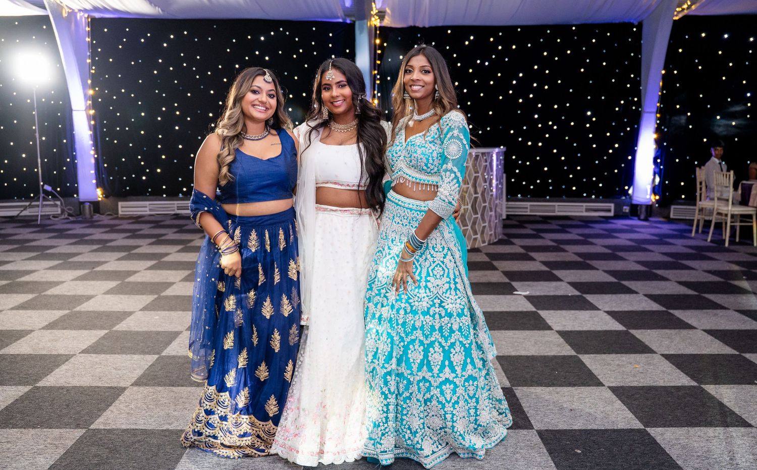 Three women in colorful Indian lehengas pose together on a black and white checkered wedding dance floor.