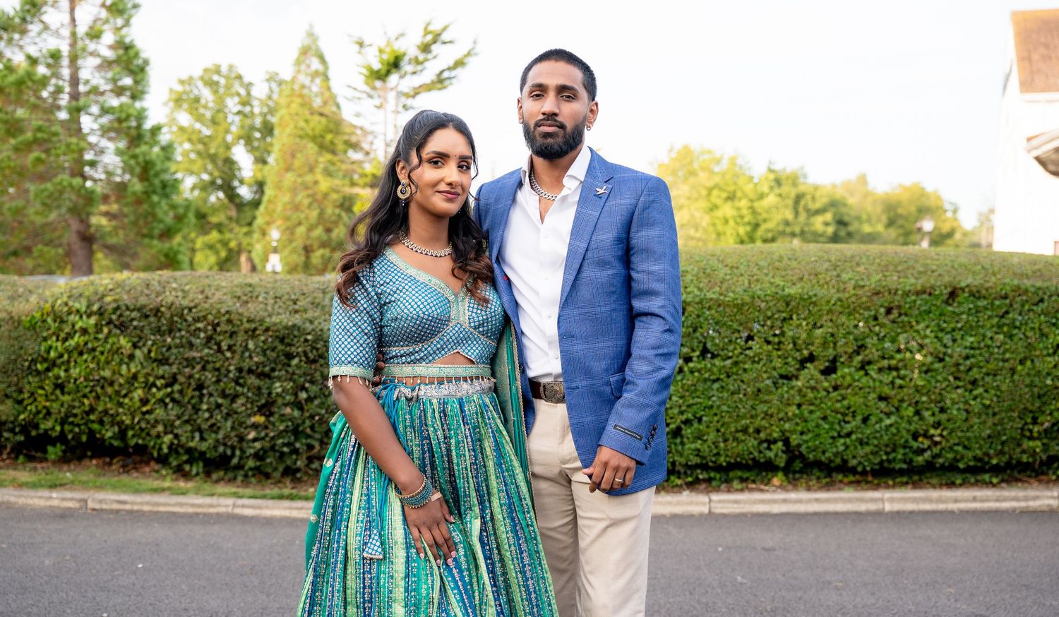 A couple poses outdoors in elegant Indian attire, the woman in a teal lehenga and the man in a blue blazer.