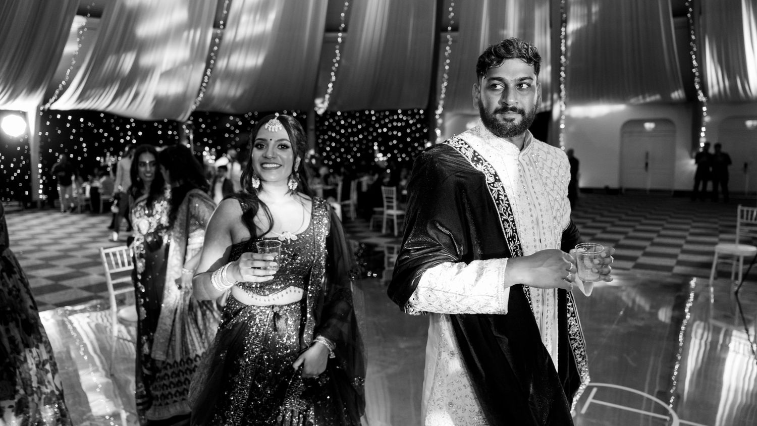 Black and white photo of bride and groom in traditional Indian attire walking at wedding reception.