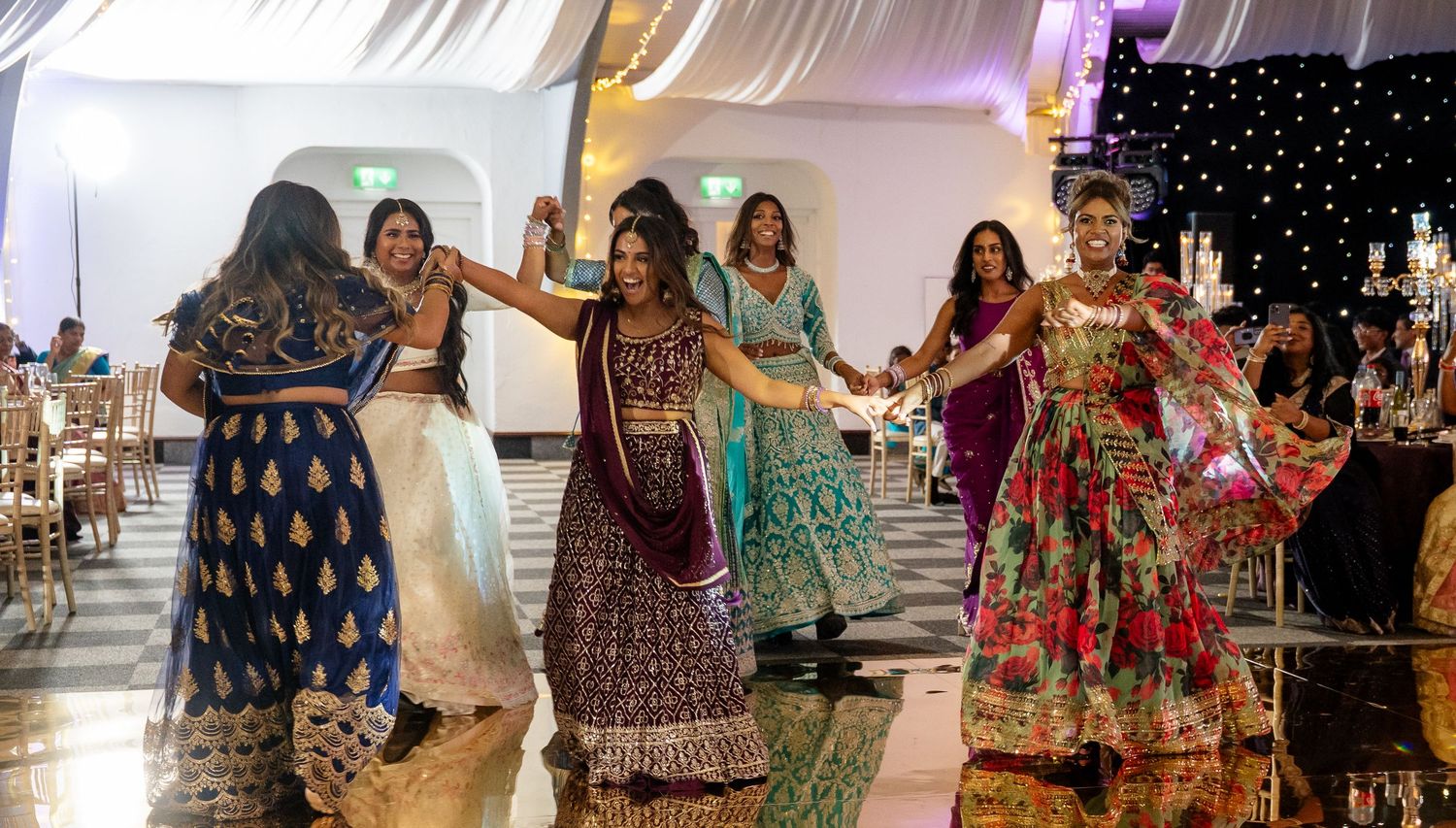 Women in colorful Indian lehengas dancing joyfully together on the dance floor at a wedding reception.
