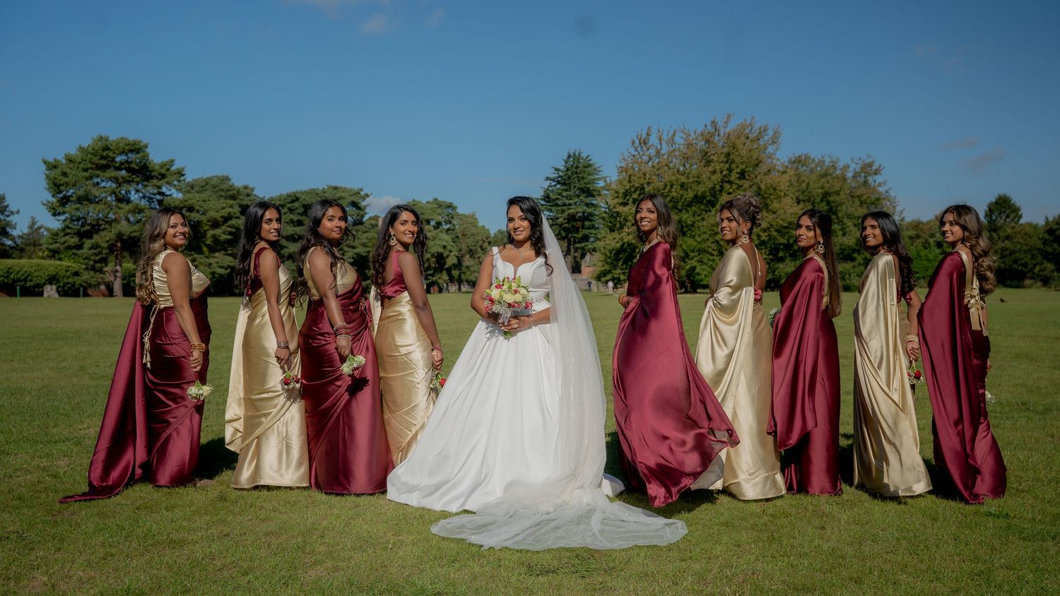 Bride in white gown surrounded by bridesmaids in burgundy and gold dresses on sunny wedding day.