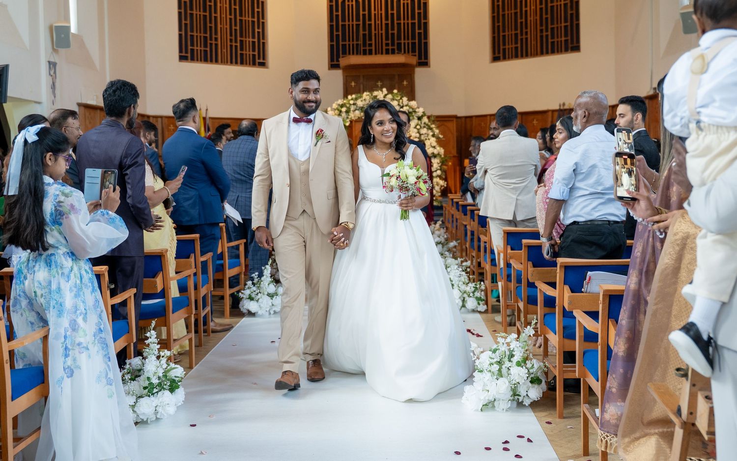 Smiling newlywed couple walks hand in hand down decorated church aisle as guests celebrate around them.