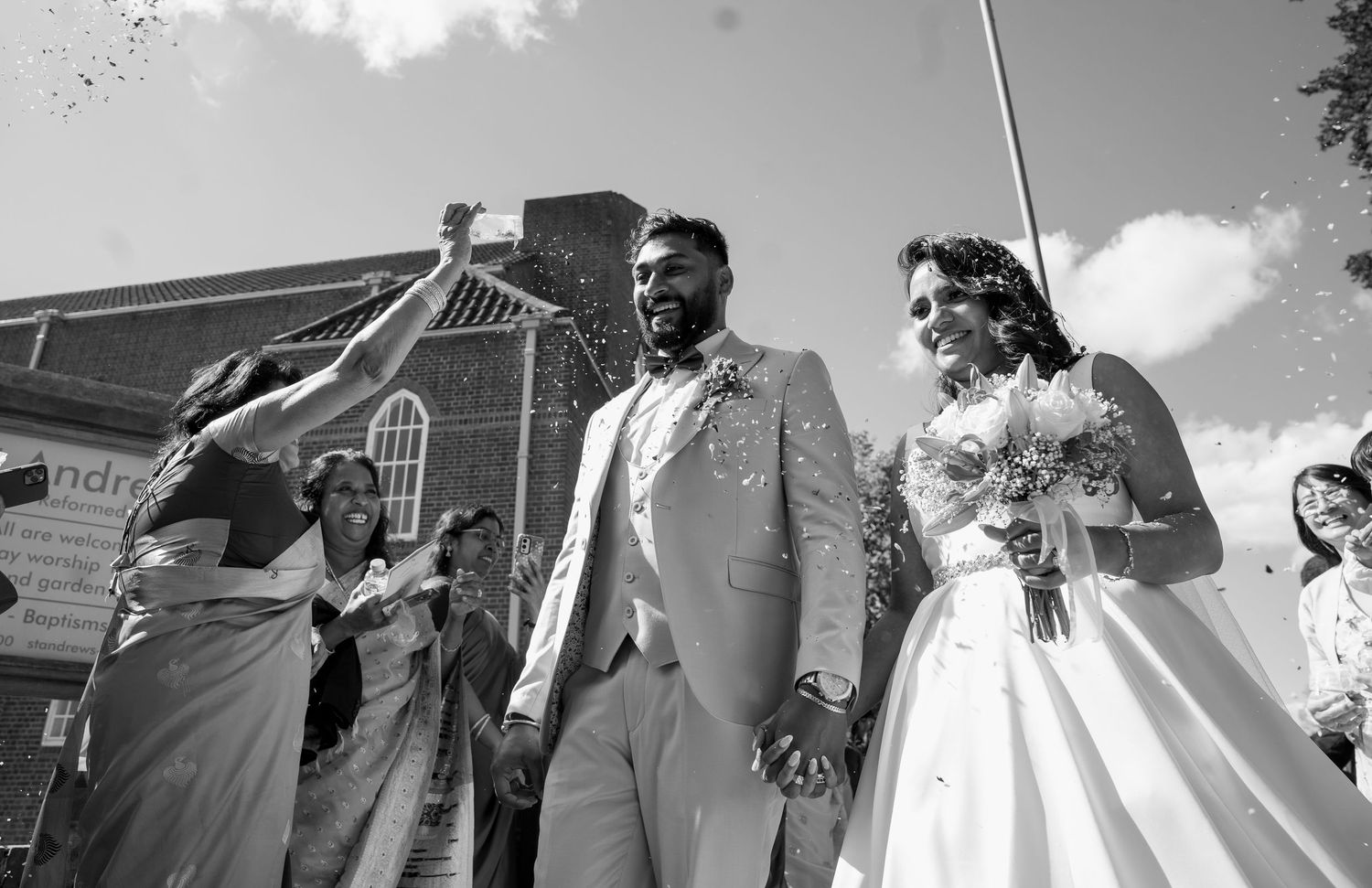 Black and white photo of newlyweds walking through a confetti shower outside a brick church.