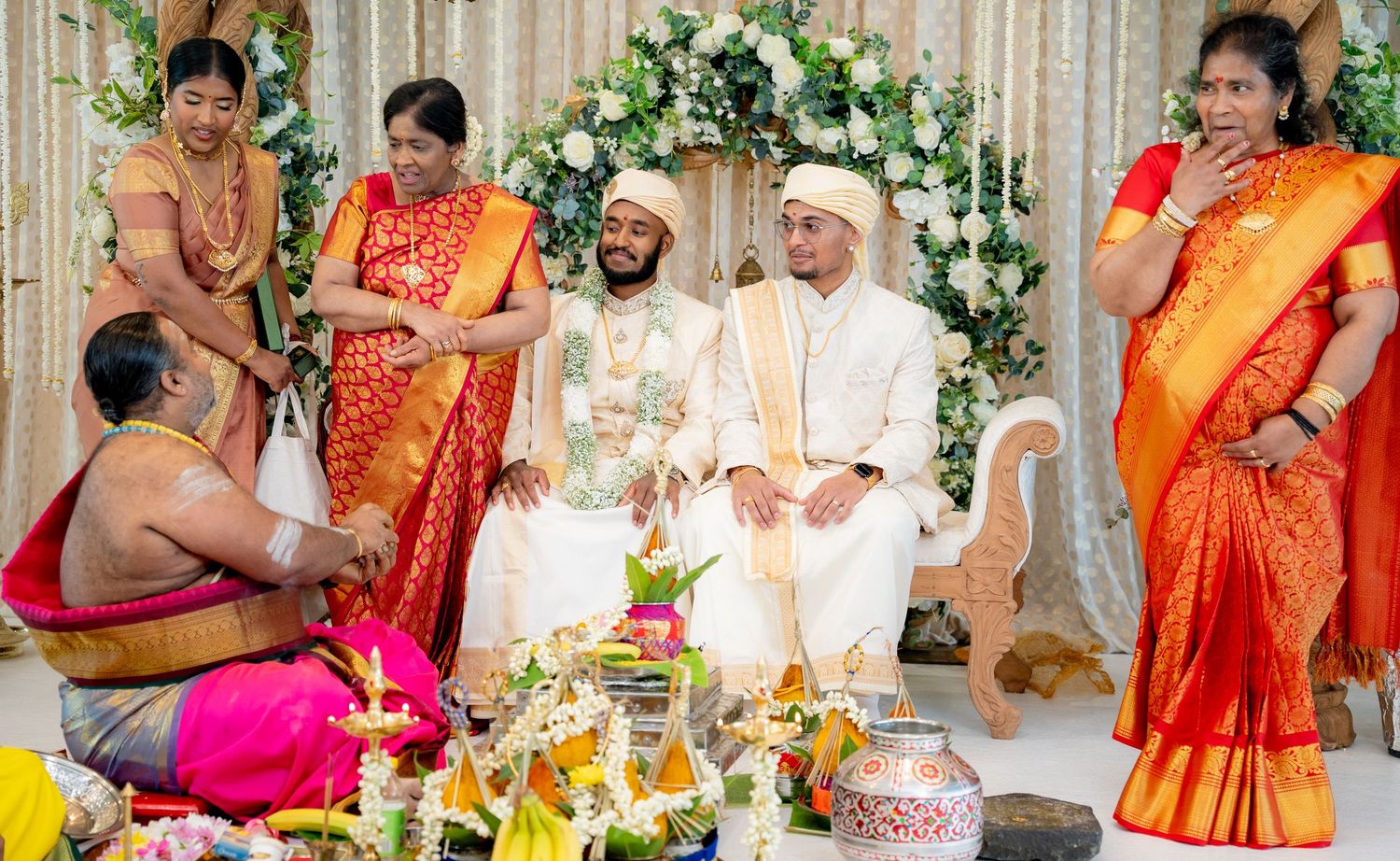 Two grooms smiling during South Indian wedding rituals surrounded by family in vibrant sarees.